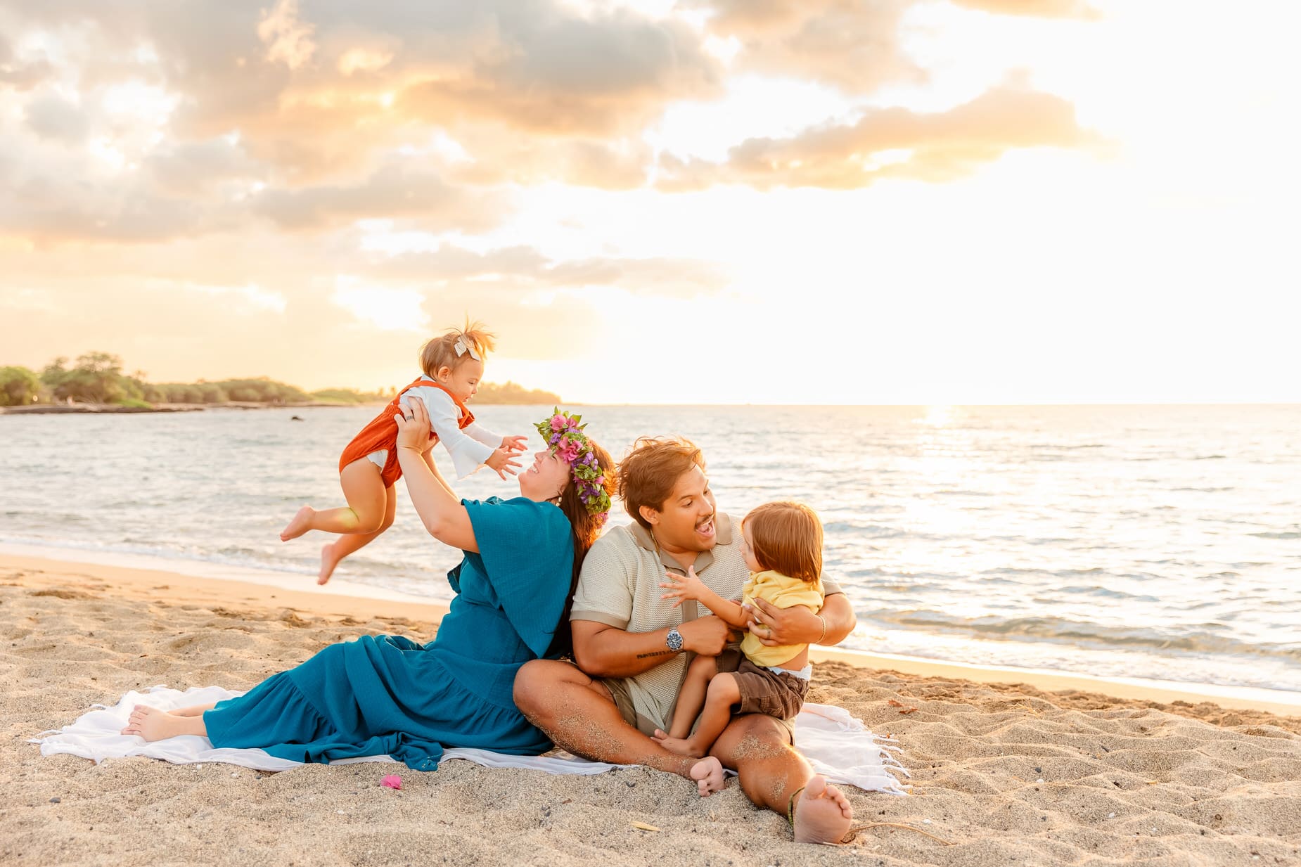 A family of four sits on a sandy beach at sunset. An adult in a blue dress lifts a toddler in the air, while another adult sits beside them holding a child on their lap. The ocean and bright sky are in the background.