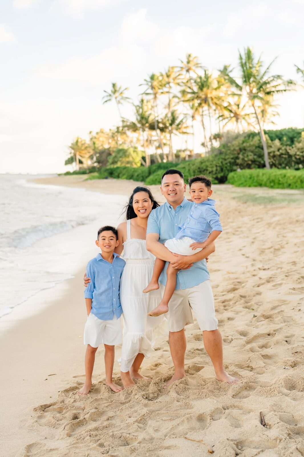 A family of four stands smiling on a sandy beach, with palm trees and greenery in the background. The parents and two young boys wear coordinated light blue and white outfits. The ocean waves are gently rolling in beside them.
