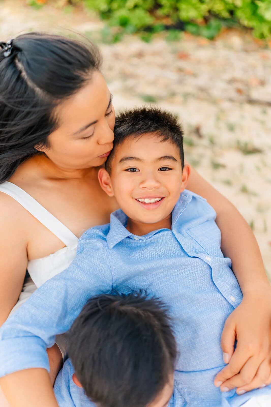A woman embraces and kisses a smiling young boy in a blue shirt while holding another child, outdoors on a sunny day with greenery in the background.