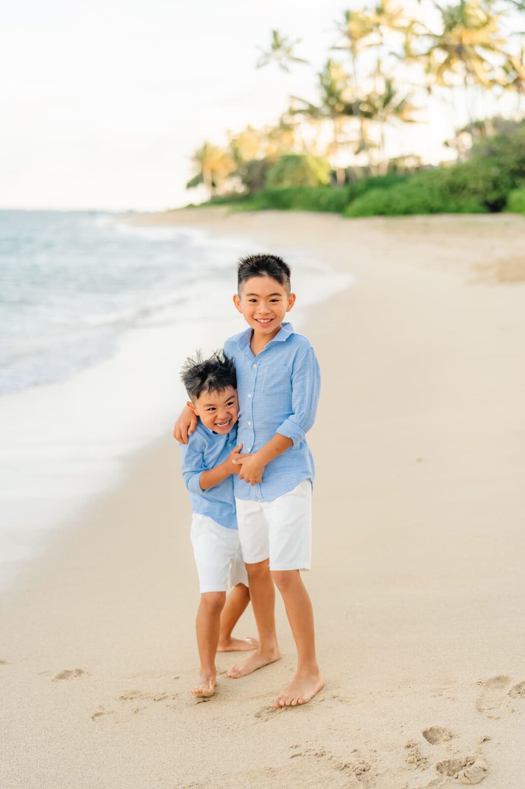 Two young boys wearing matching blue shirts and white shorts smile and hug each other while standing barefoot on a sandy beach, with ocean waves and palm trees in the background.