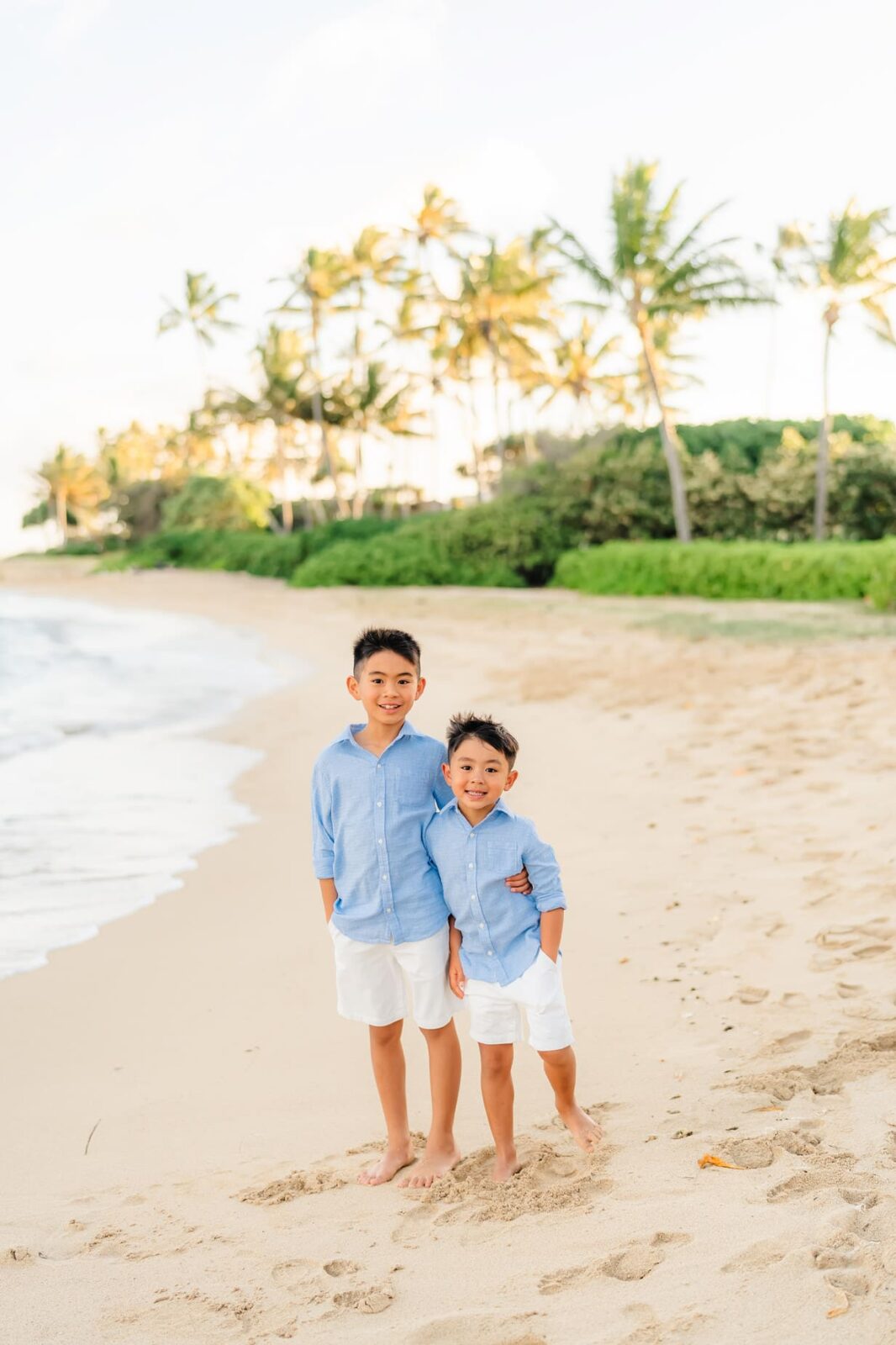 Two young boys in matching light blue shirts and white shorts stand barefoot on a sandy beach, smiling and hugging. Palm trees and green bushes line the background under a bright sky.
