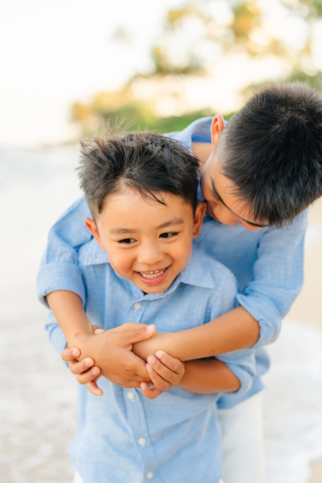 Two young boys wearing light blue shirts smile and hug each other on a beach, with soft sunlight and palm trees blurred in the background. The older boy embraces the younger from behind.