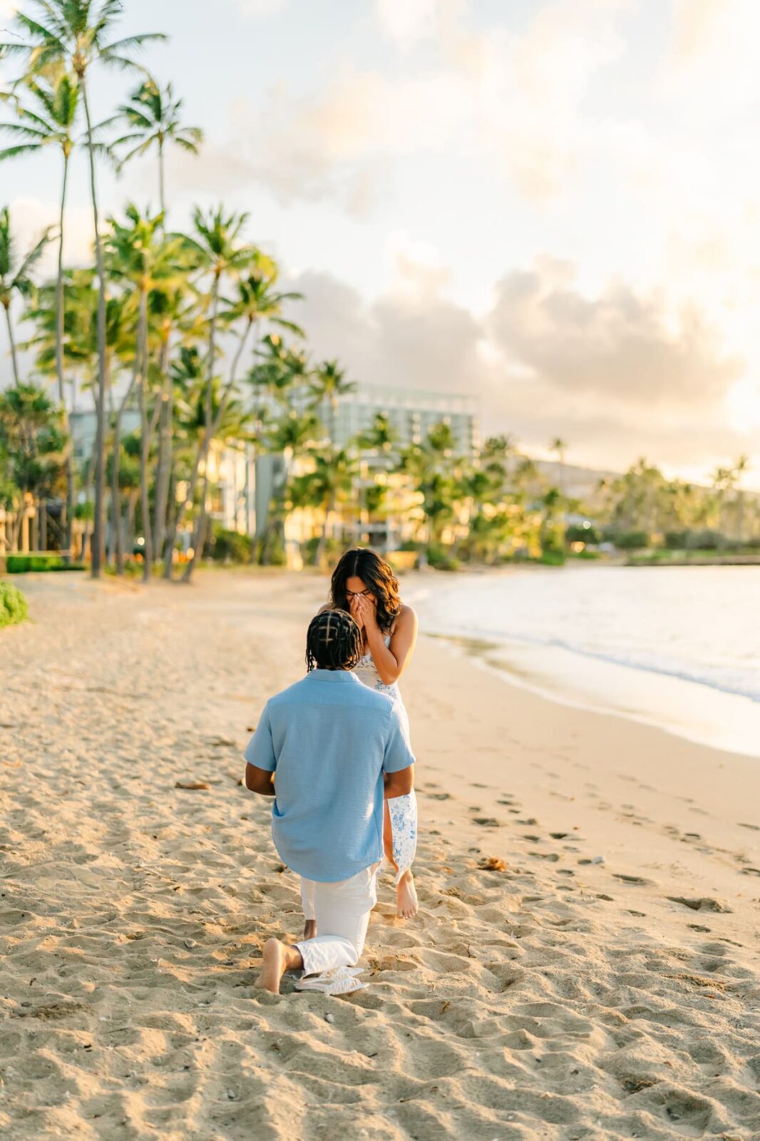 A man kneels on a sandy beach at sunset, proposing to a woman who covers her mouth in surprise—a perfect Hawaii surprise proposal framed by palm trees and gentle waves in the background.