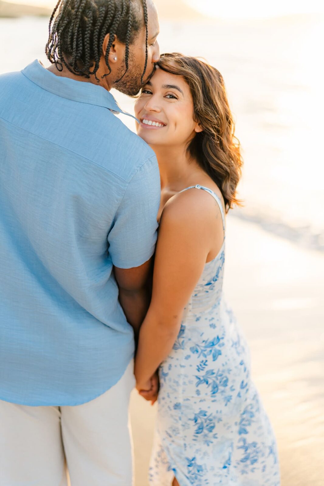 A man kisses a smiling woman on the forehead as they hold hands on a sunlit beach during their Hawaii surprise proposal. She wears a white and blue floral dress and looks at the camera, while he sports a light blue shirt and light-colored pants.