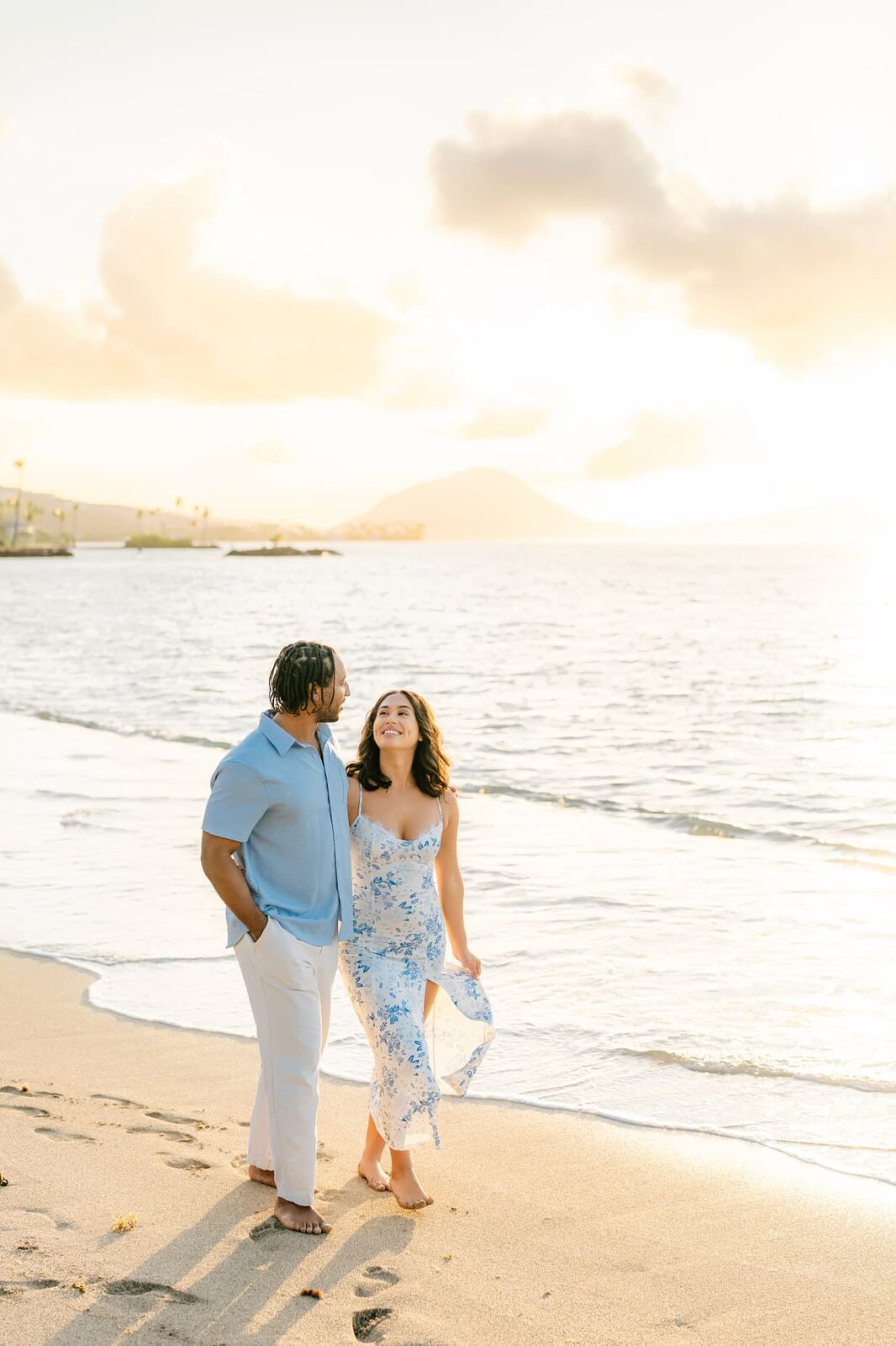 A couple walks barefoot along a sandy beach at sunset in Hawaii, smiling at each other during a surprise proposal. The ocean and distant mountains are visible under a bright, golden sky.