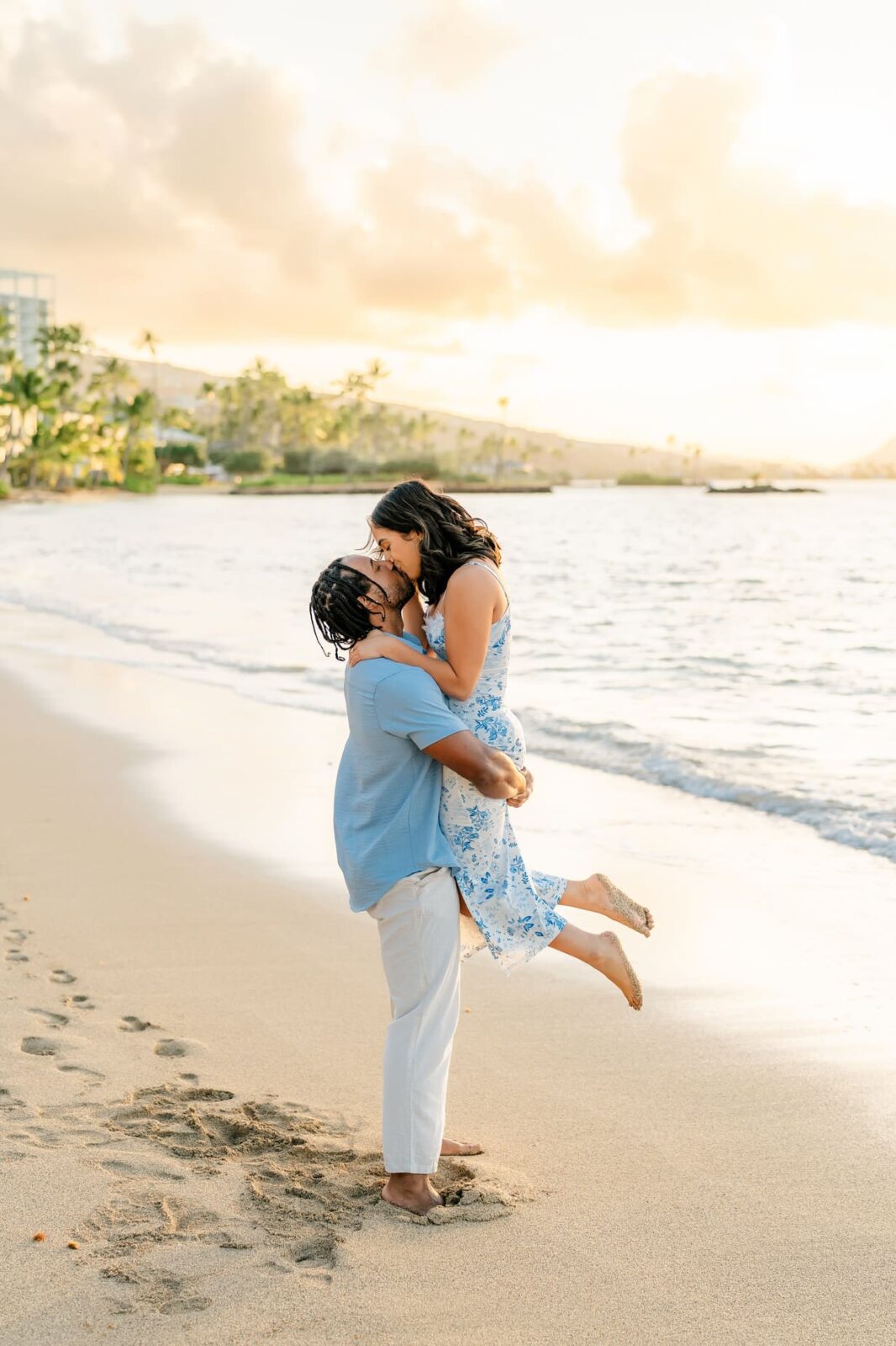 A man lifts a woman in a blue floral dress as they kiss on a sandy beach at sunset, with ocean waves and palm trees behind them. Both, barefoot and joyful, share a romantic Hawaii surprise proposal moment.