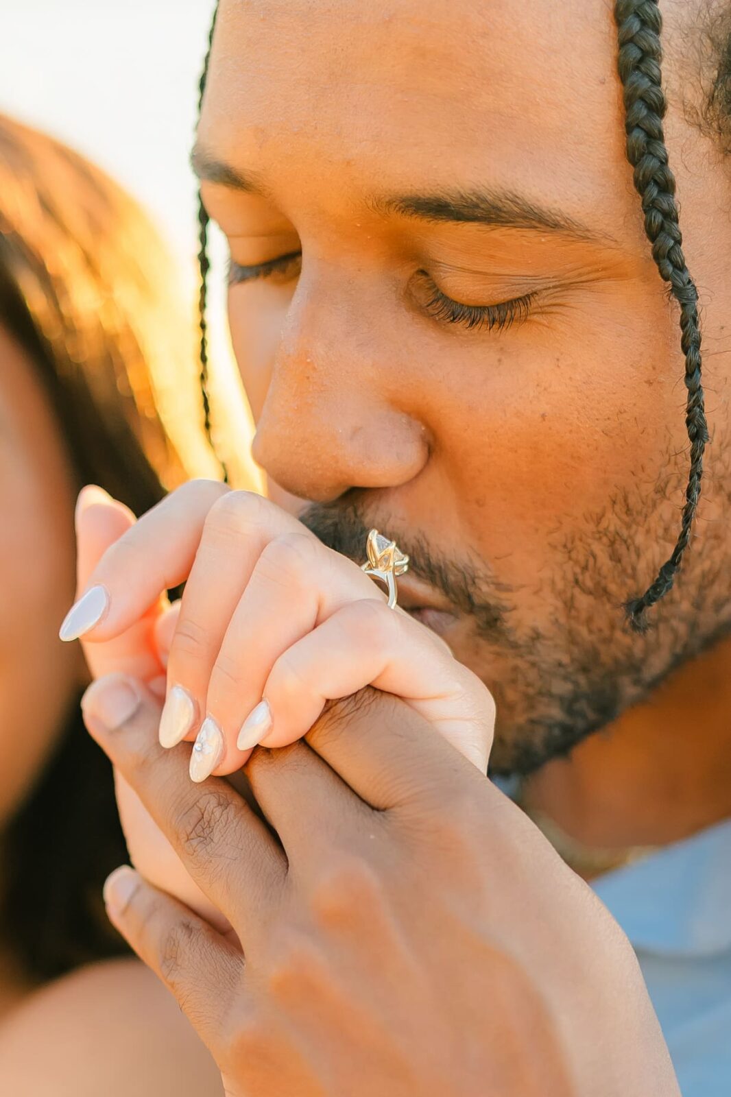 A man with braids gently kisses a woman’s hand, revealing her engagement ring in this Hawaii surprise proposal. The close-up shot highlights tenderness and affection between the couple, with warm sunlight in the background.
