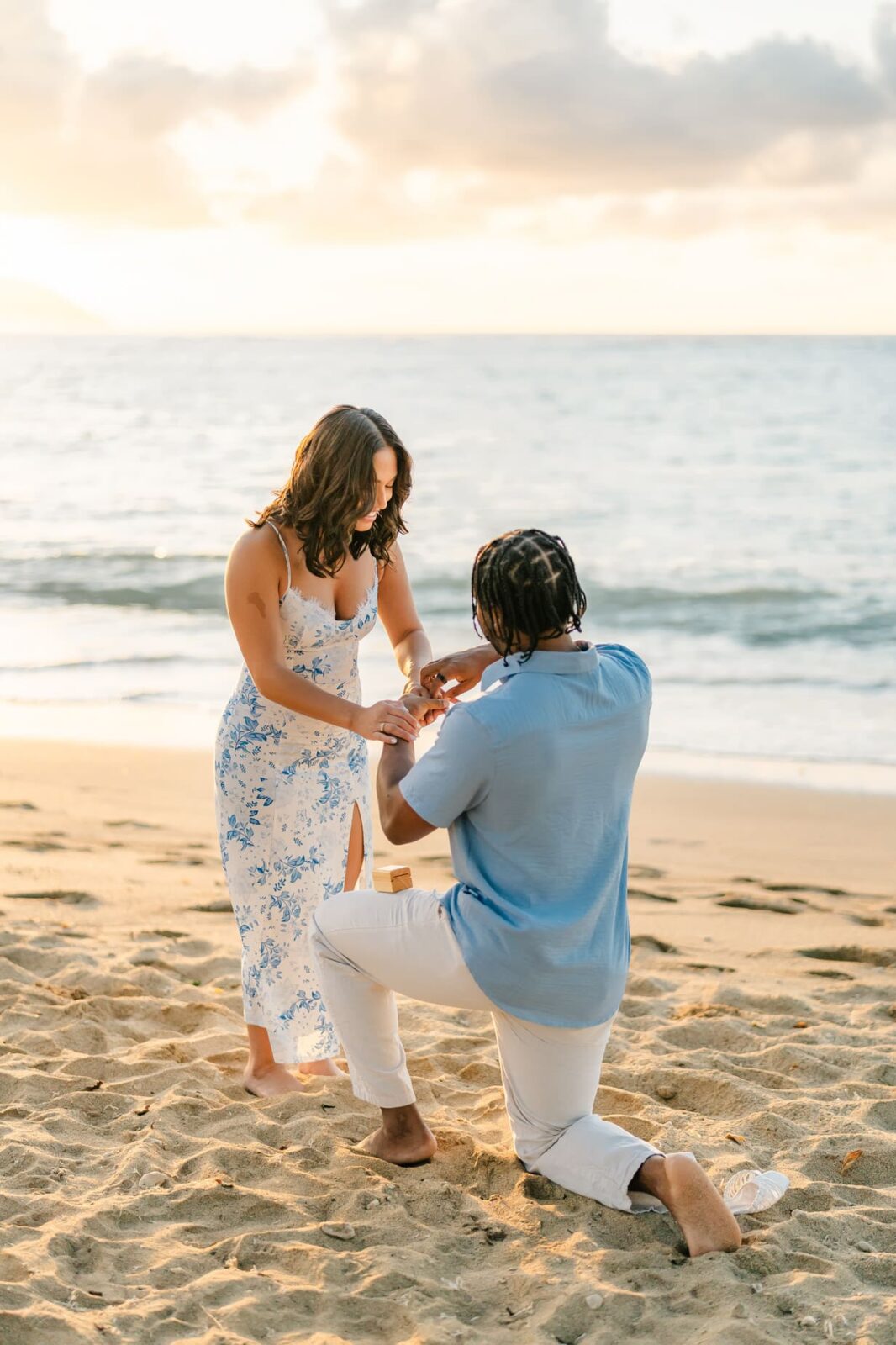 A man kneels on one knee, making a Hawaii surprise proposal to a woman on a sandy beach at sunset. She wears a white and blue dress, and he wears a light blue shirt with white pants. The ocean and sky create a stunning backdrop.