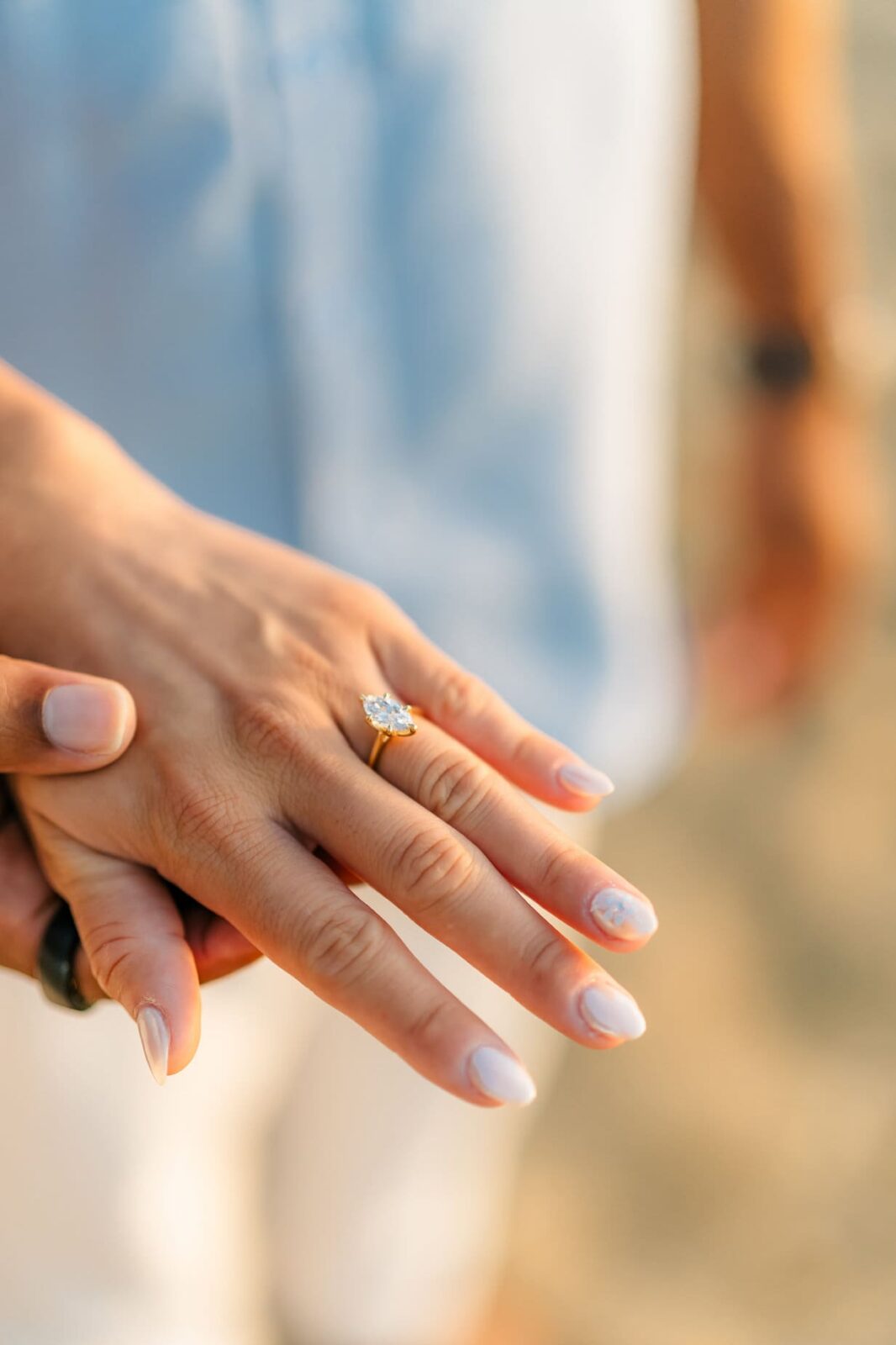 A close-up of a hand with pale pink manicured nails wearing an engagement ring with a large diamond, gently held by another person during a Hawaii surprise proposal. The background is softly blurred.