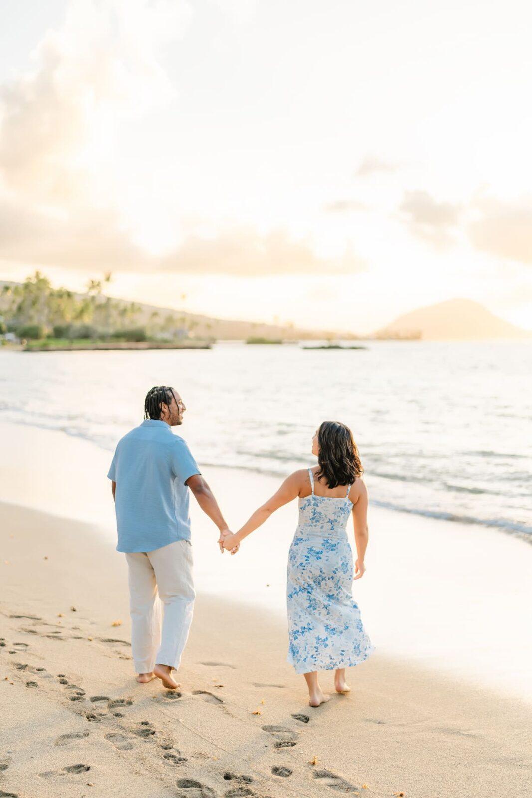 A couple holding hands walks barefoot along a sandy beach at sunset, leaving footprints behind—perfect for a Hawaii surprise proposal. The woman wears a blue floral dress, the man a light blue shirt and white pants. Ocean and hills complete the scene.