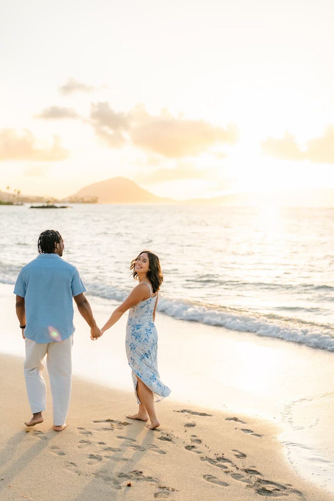 A couple holds hands and walks along a sandy beach at sunset in Hawaii. The woman, wearing a light blue floral dress, smiles back, unaware of the surprise proposal the man in a light blue shirt and white pants has planned. Waves and mountains complete the scene.