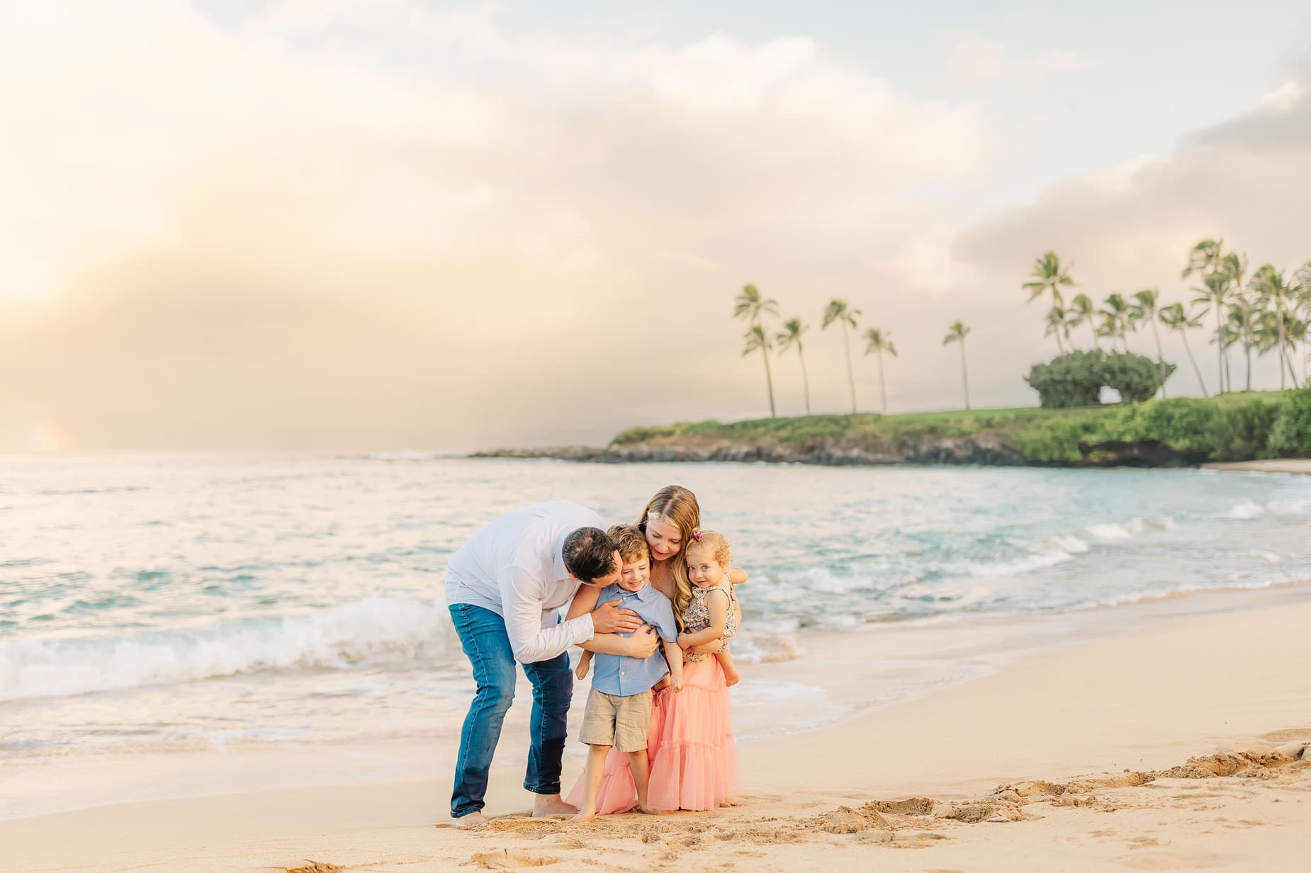 A family of four hugs on a sandy beach at sunset, with waves in the background and palm trees along the shoreline. The parents lean in close to their two young children, smiling and enjoying a magical Kapalua Sunrise together.