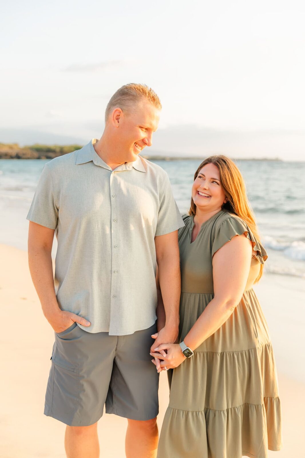 A smiling couple holds hands and looks at each other while standing on a sandy beach near the ocean, with sunlight illuminating them.