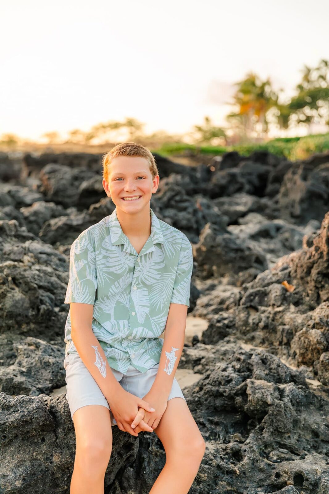 A smiling boy in a green tropical shirt and light shorts sits on black volcanic rocks outdoors, with palm trees and a bright sky in the background.