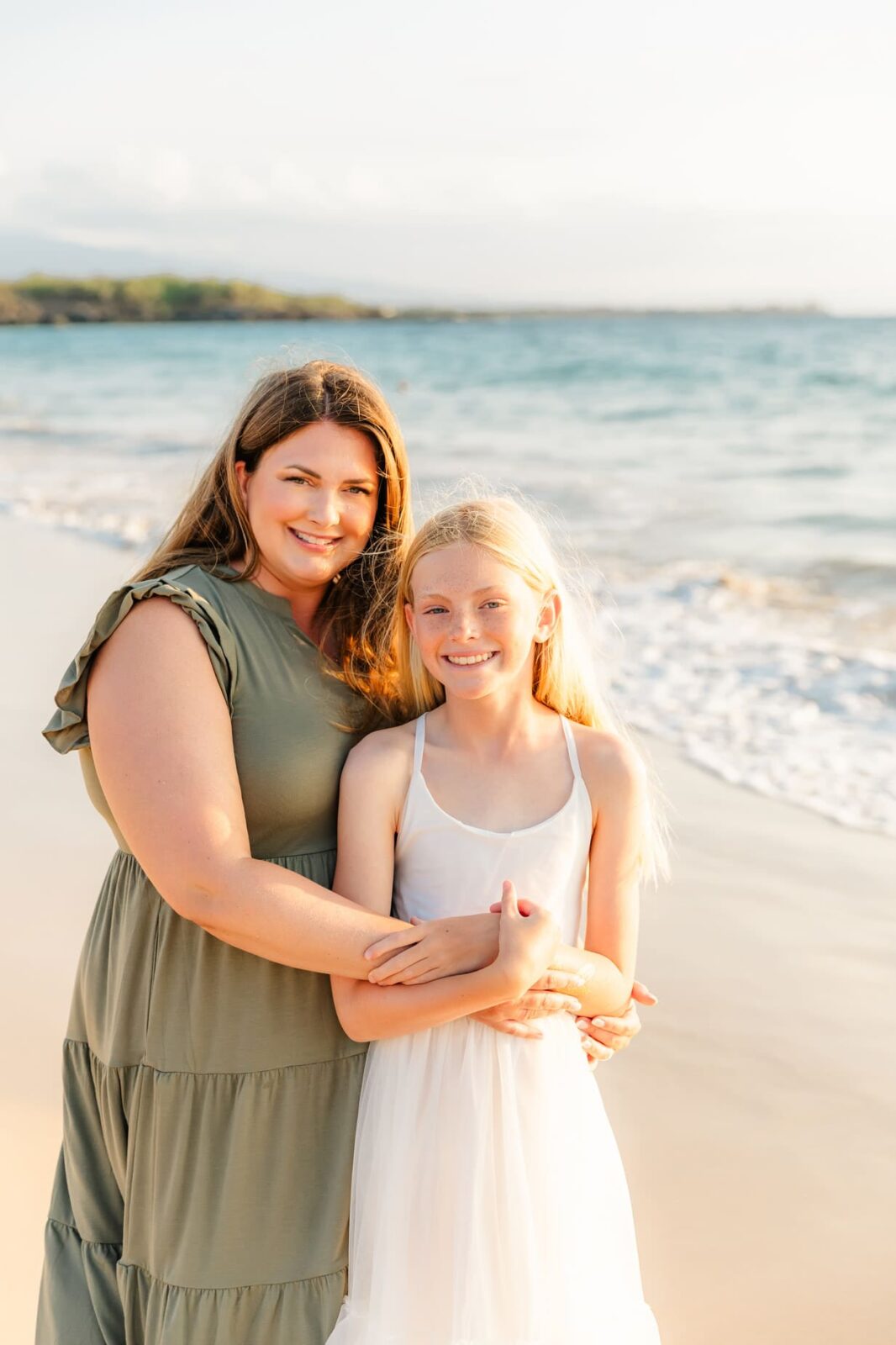 A woman in a green dress hugs a young girl in a white dress as they smile on a sandy beach with gentle waves and sunlight in the background.
