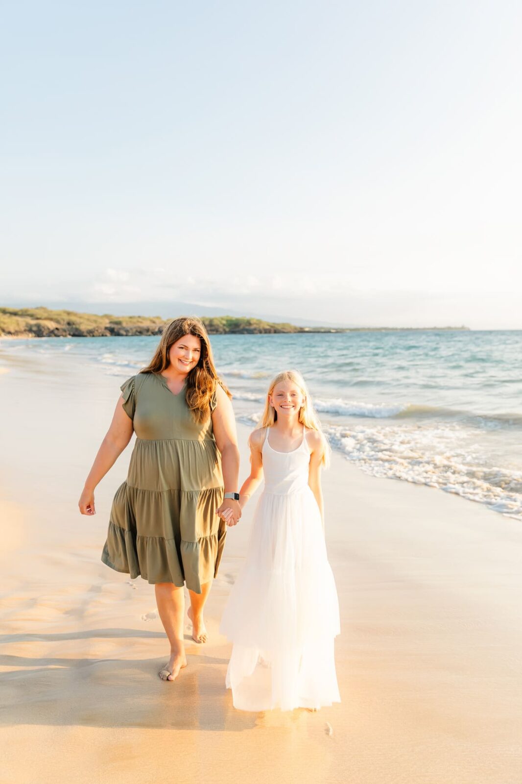 A woman in a green dress and a young girl in a white dress walk hand-in-hand along a sandy beach, smiling, with gentle waves and sunlight in the background.