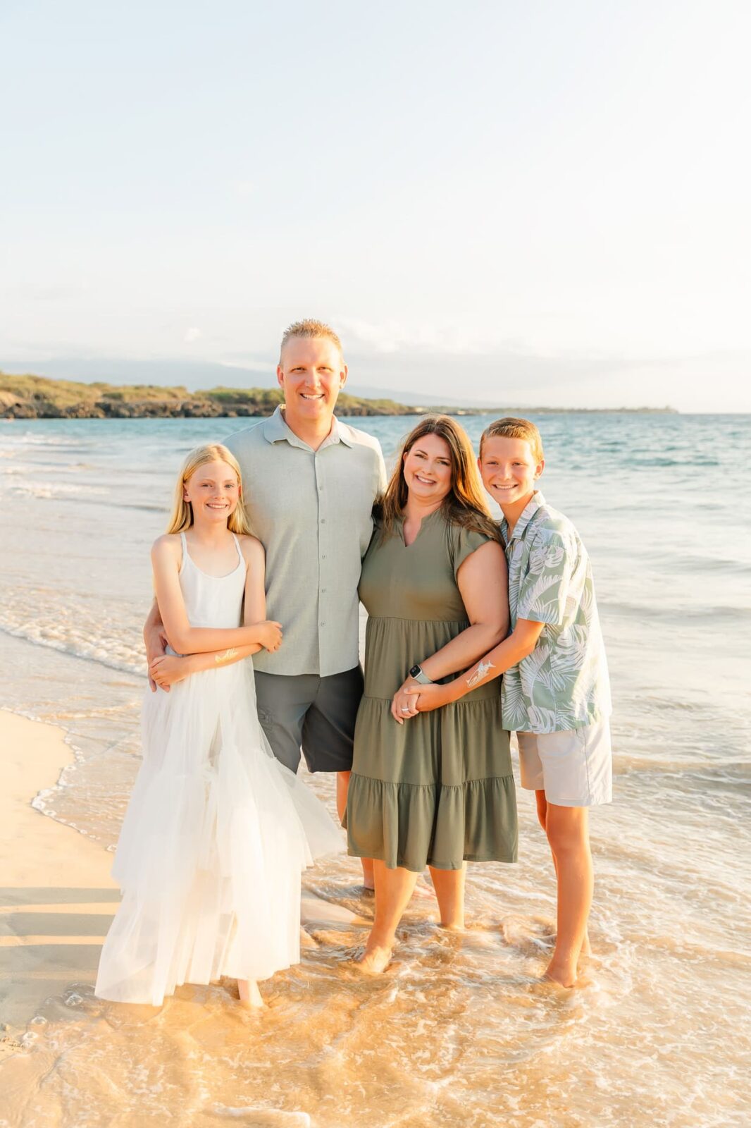 A family of four stands smiling on a sandy beach at sunset. The parents are in the center, while a girl in a white dress and a boy in a tropical shirt stand on either side. Gentle waves and coastline are in the background.