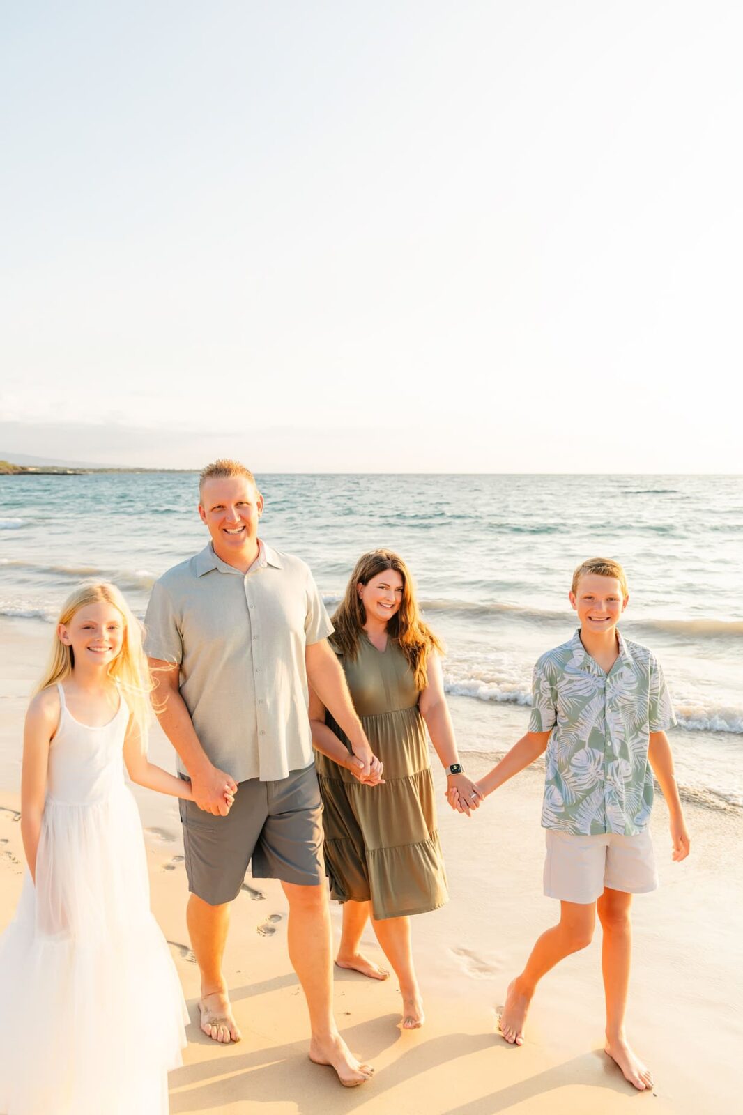 A smiling family of four walks hand-in-hand along a sunny beach. The mother and father are in the center, with a daughter in a white dress and a son in a patterned shirt and shorts on either side. The ocean is in the background.