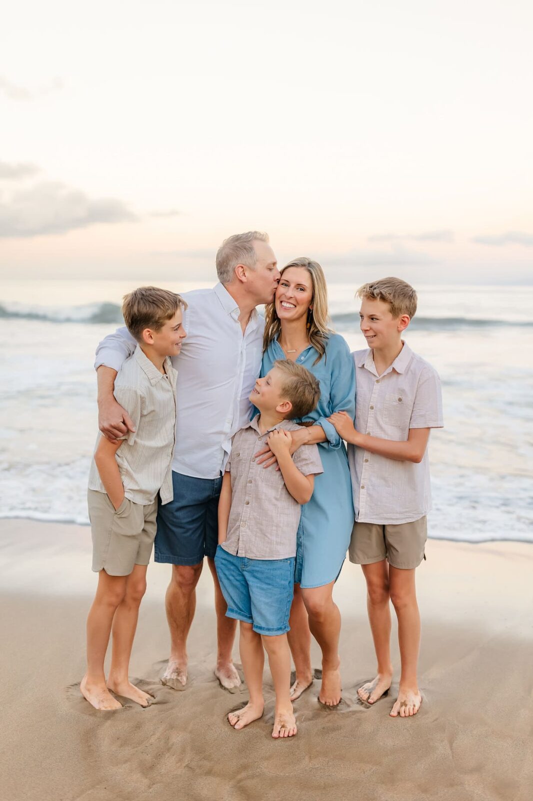 A family of five stands barefoot on the beach, smiling and laughing together. The father kisses the mother on the cheek, while their three sons stand close, with waves and a pastel sky in the background.