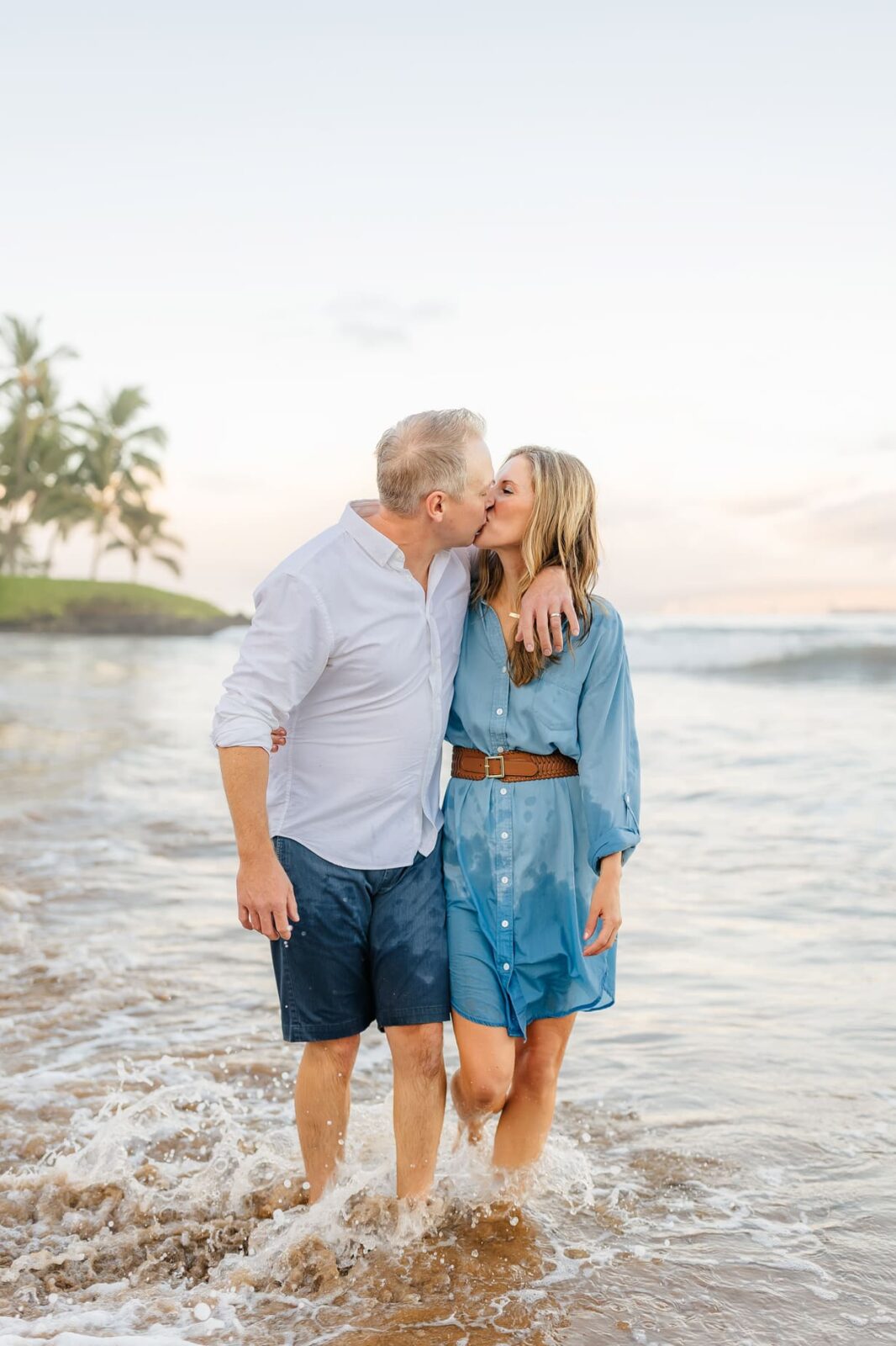 A couple stands in shallow ocean water at the beach, kissing and smiling. Both are dressed casually; the man in a white shirt and navy shorts, and the woman in a blue dress. Palm trees and a calm sky are in the background.