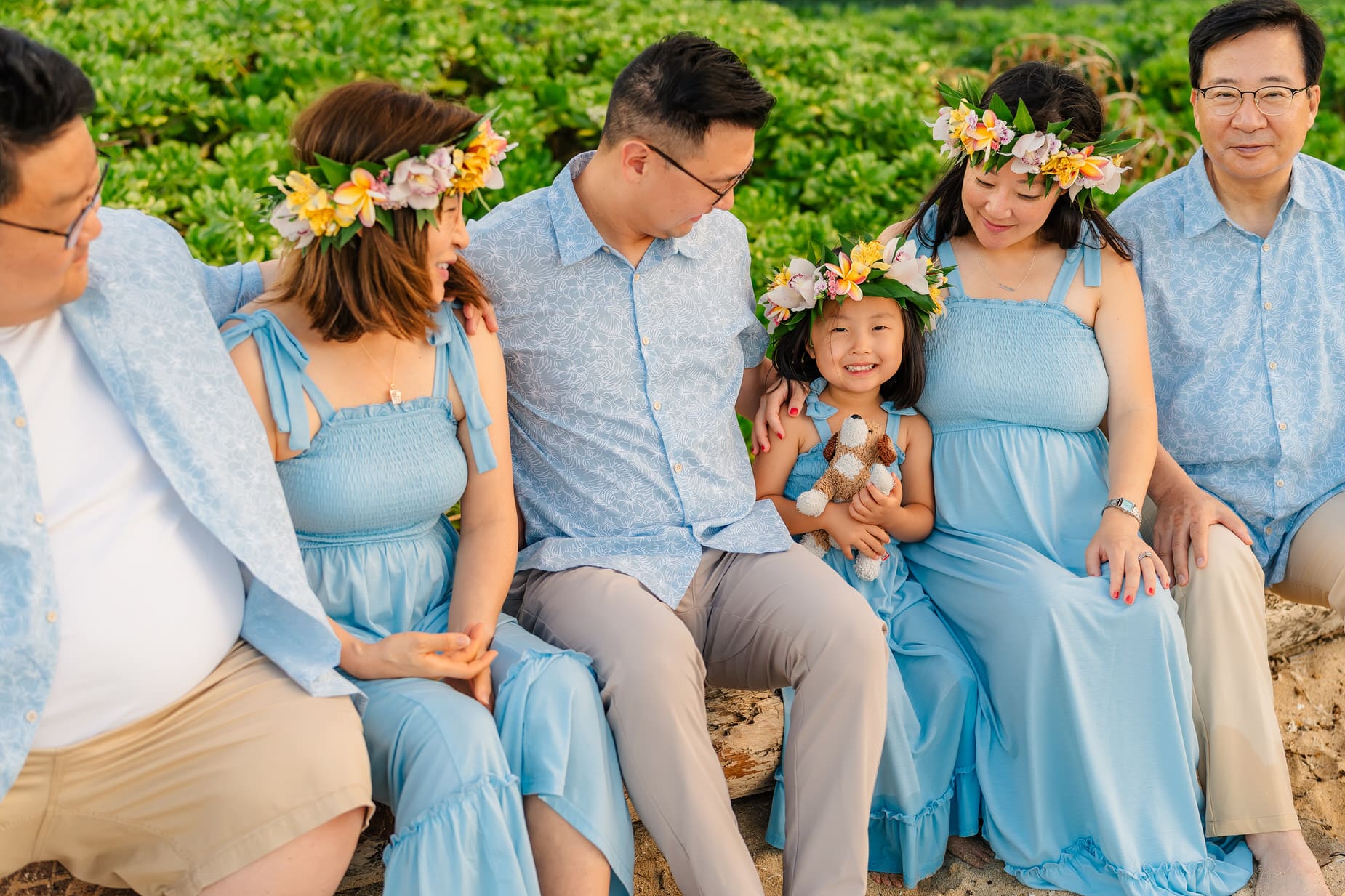 A family sits together outdoors on a log, wearing coordinated blue outfits and floral crowns. A young girl in the center smiles, holding a stuffed animal, surrounded by adults. Green foliage is in the background.