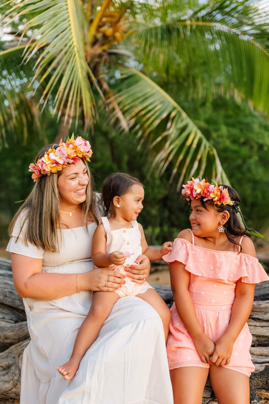 A woman and two young girls wearing floral crowns and light dresses sit together on a log, smiling and laughing, with a palm tree and lush greenery in the background.