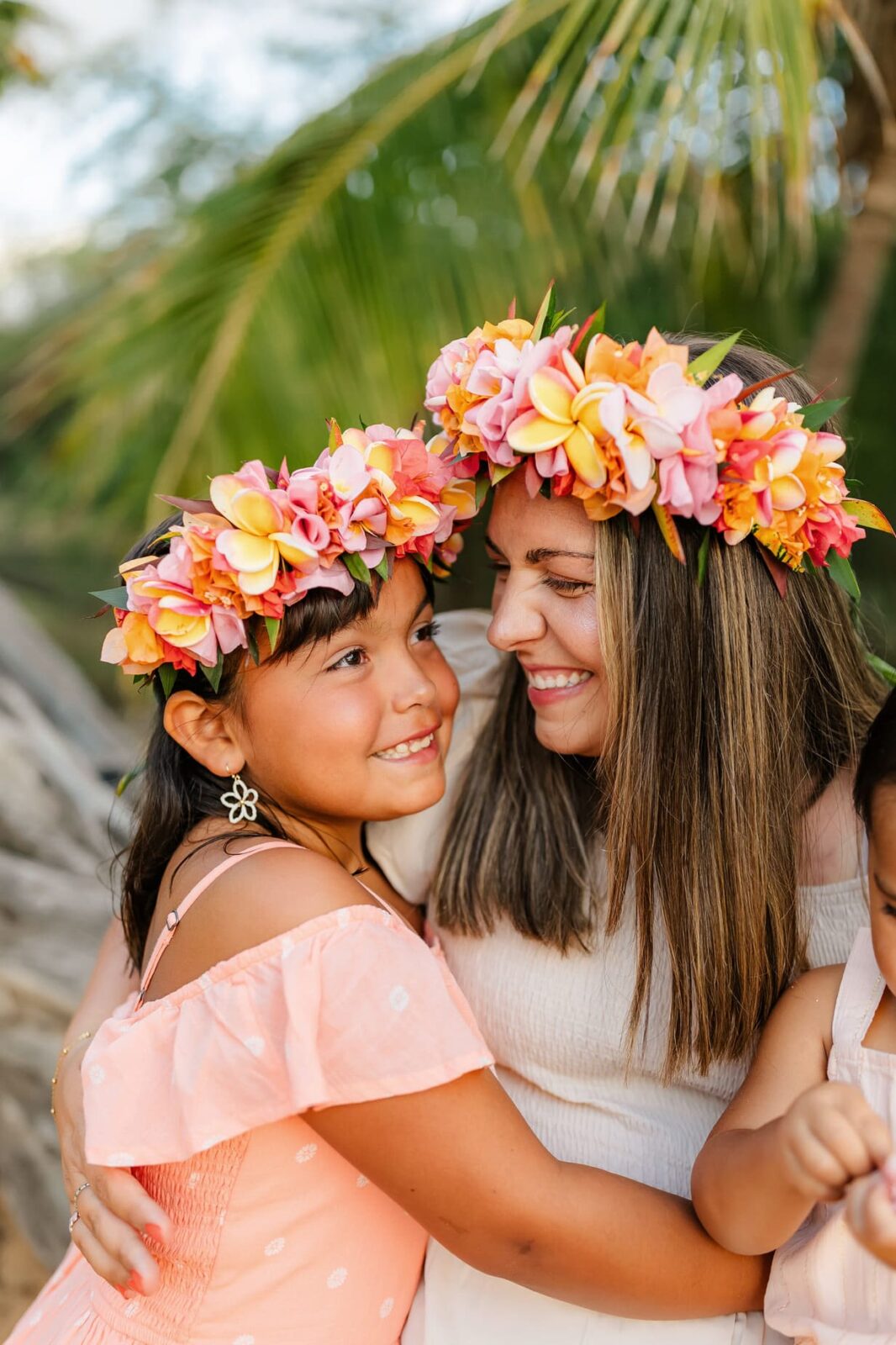 A smiling woman and girl wearing flower crowns embrace outdoors, both dressed in peach-toned outfits, with greenery and palm leaves in the background.