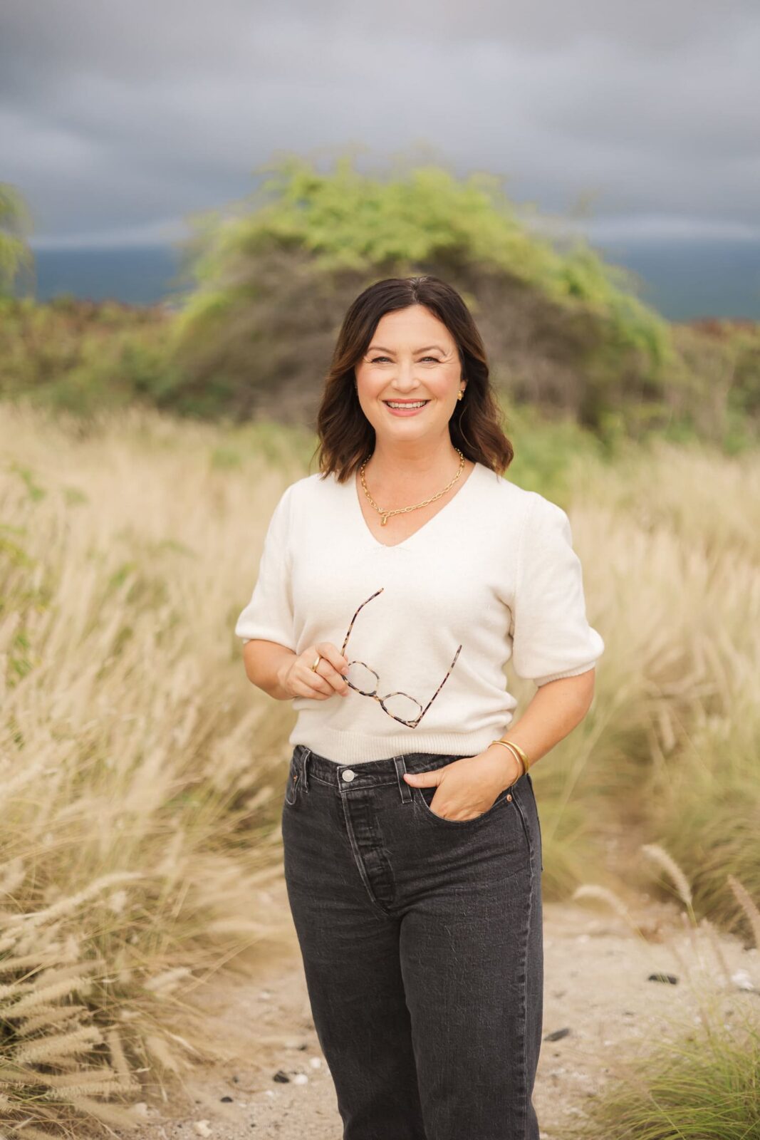 A woman with shoulder-length dark hair, wearing a light short-sleeve sweater and dark jeans, stands outdoors in tall grass, holding eyeglasses and smiling at the camera. Green foliage and a cloudy sky are in the background.