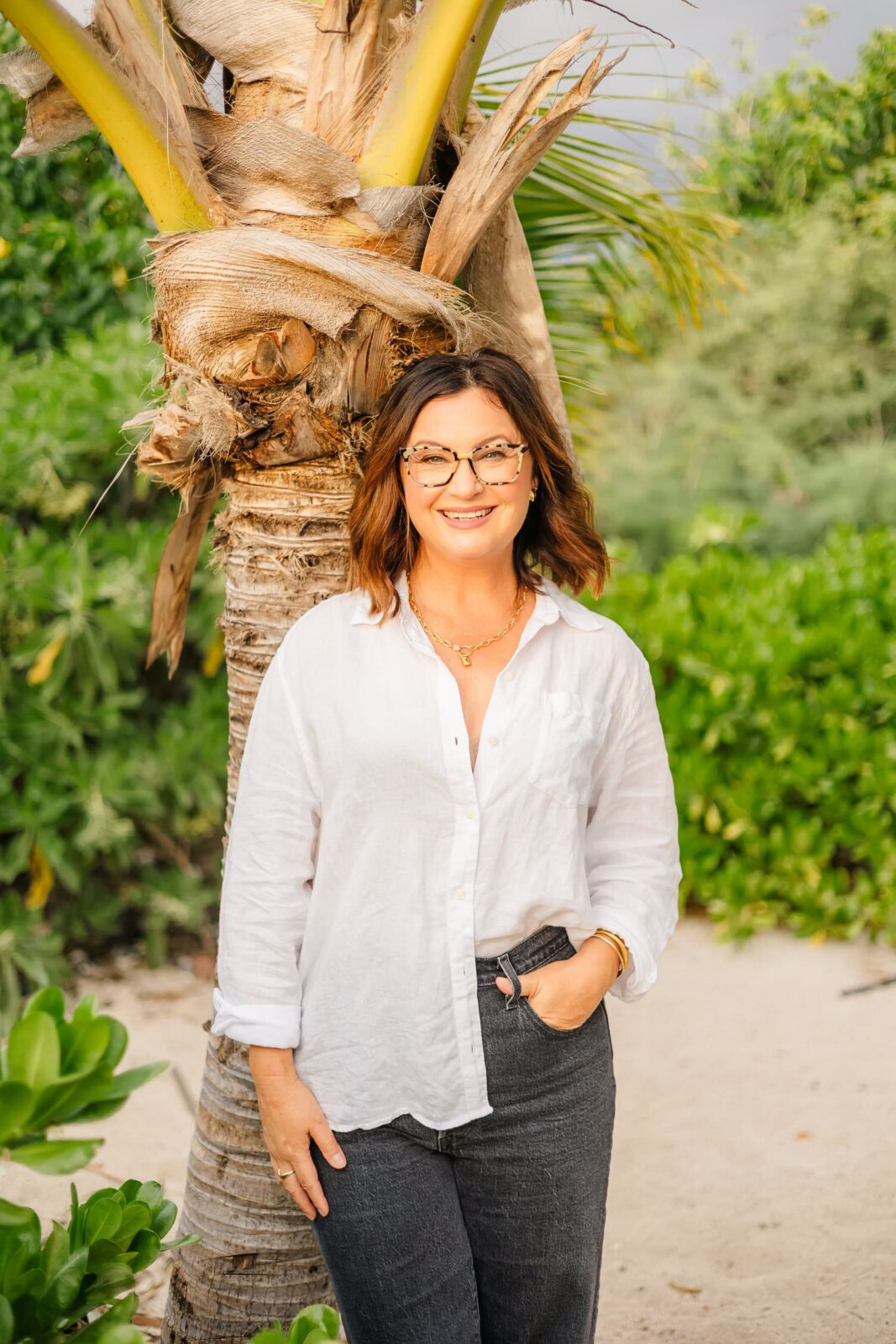 A woman wearing glasses, a white button-up shirt, and dark pants stands smiling with one hand in her pocket in front of a palm tree, surrounded by lush greenery and sandy ground.