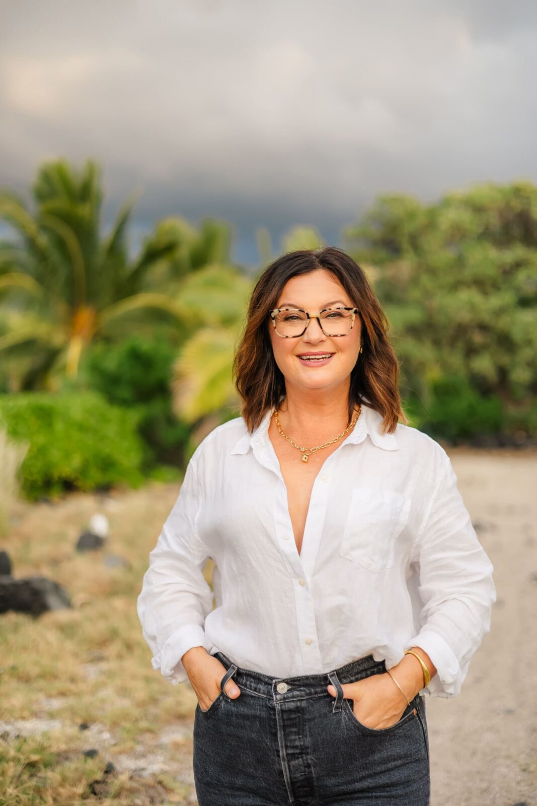 A woman with shoulder-length brown hair and glasses stands outdoors, smiling, wearing a white button-down shirt and dark jeans, with her hands in her pockets. Lush greenery and palm trees are in the background.