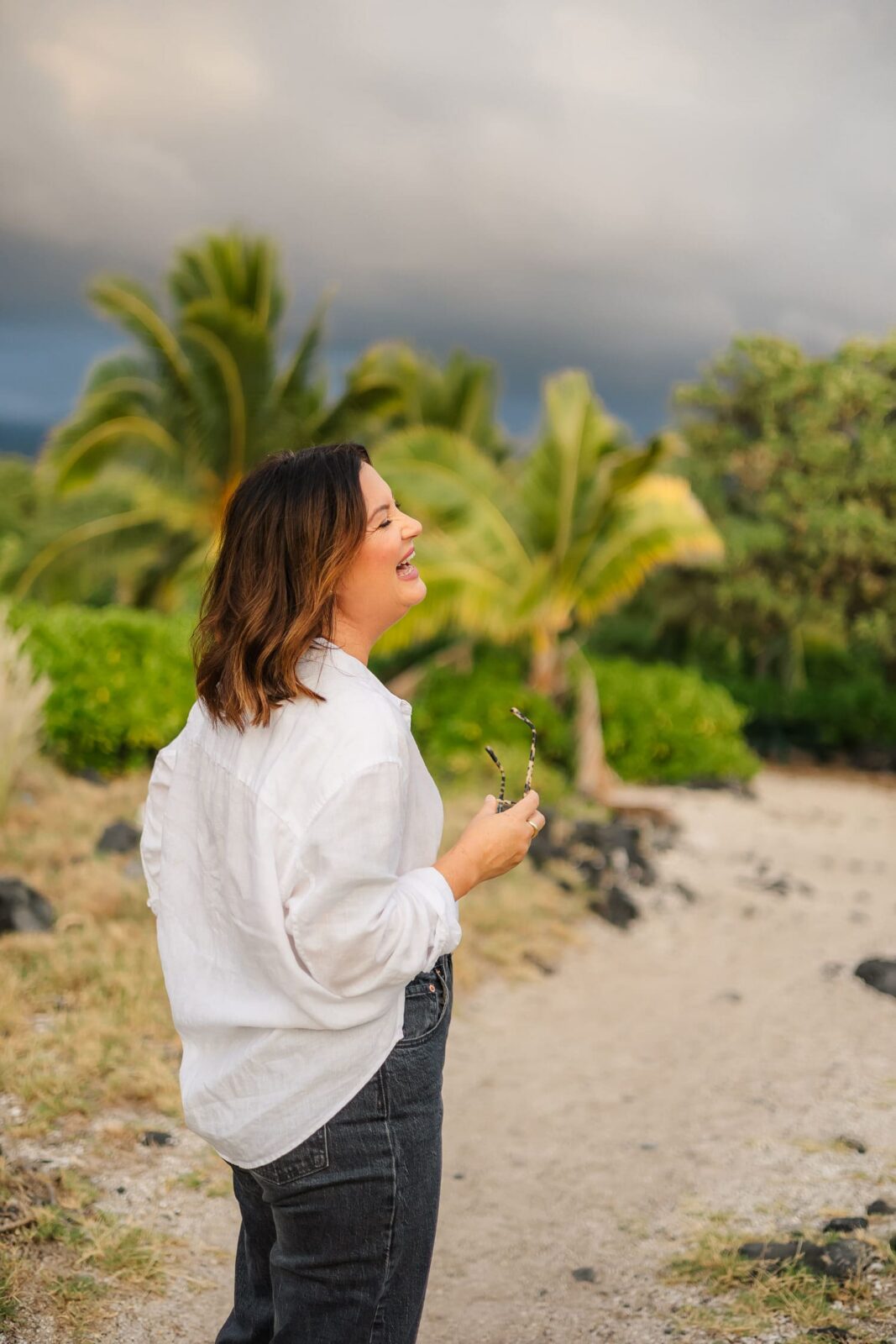 A woman in a white shirt and dark jeans smiles while standing on a sandy path surrounded by greenery and palm trees, holding sunglasses in one hand. The sky appears cloudy in the background.