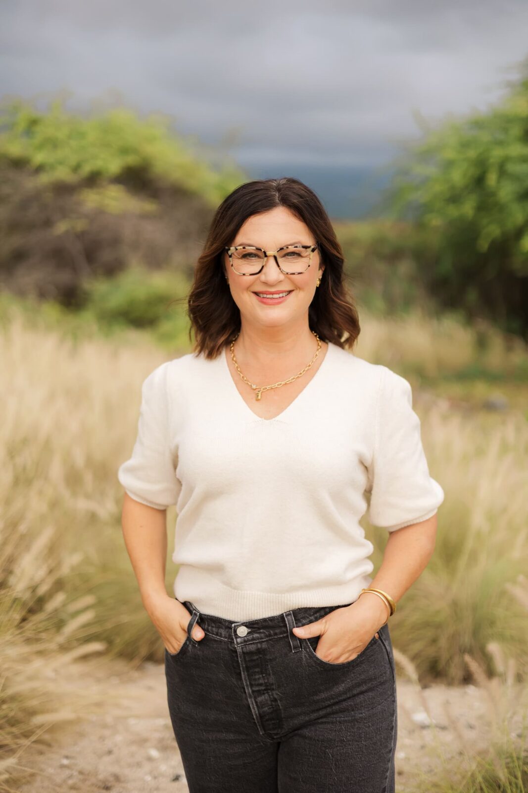 A woman with shoulder-length brown hair and glasses stands outdoors in a grassy area, wearing a white short-sleeve sweater, dark jeans, and gold jewelry, smiling with her hands in her pockets. Trees and cloudy sky are in the background.