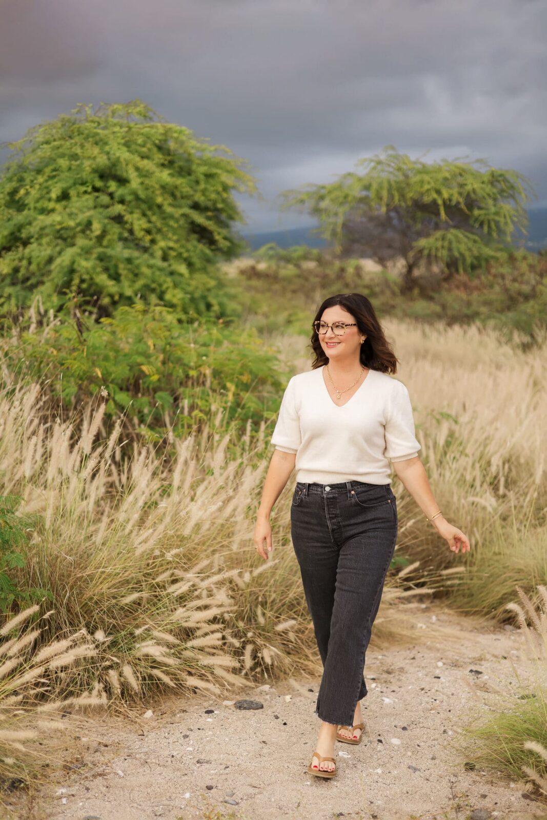 A woman with dark hair and glasses walks along a sandy path surrounded by tall grass and green bushes under a cloudy sky. She is wearing a white top, dark jeans, and sandals.