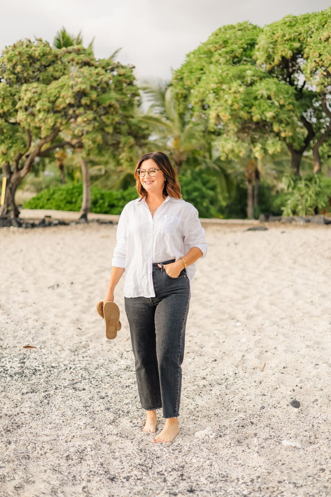 A woman with shoulder-length brown hair, wearing glasses, a white shirt, and black jeans, walks barefoot on a sandy beach, holding sandals in one hand, with green trees and plants in the background.