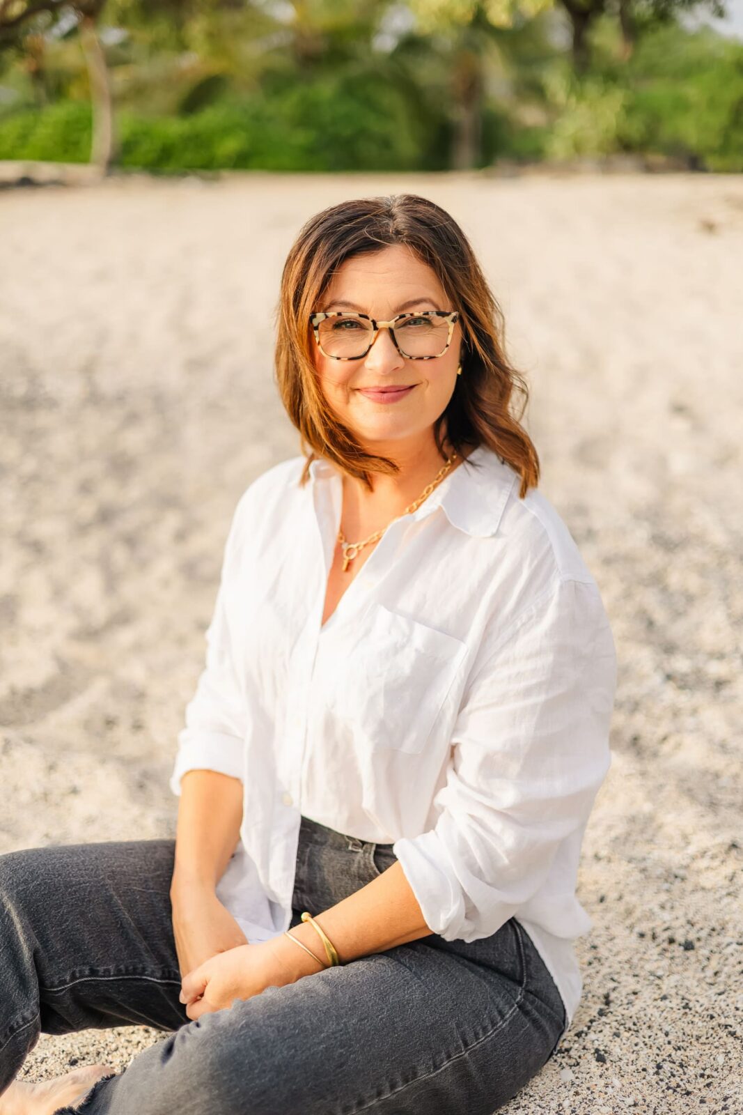 A woman with shoulder-length brown hair, wearing glasses, a white button-up shirt, and dark jeans, sits on a sandy beach, smiling at the camera with greenery blurred in the background.