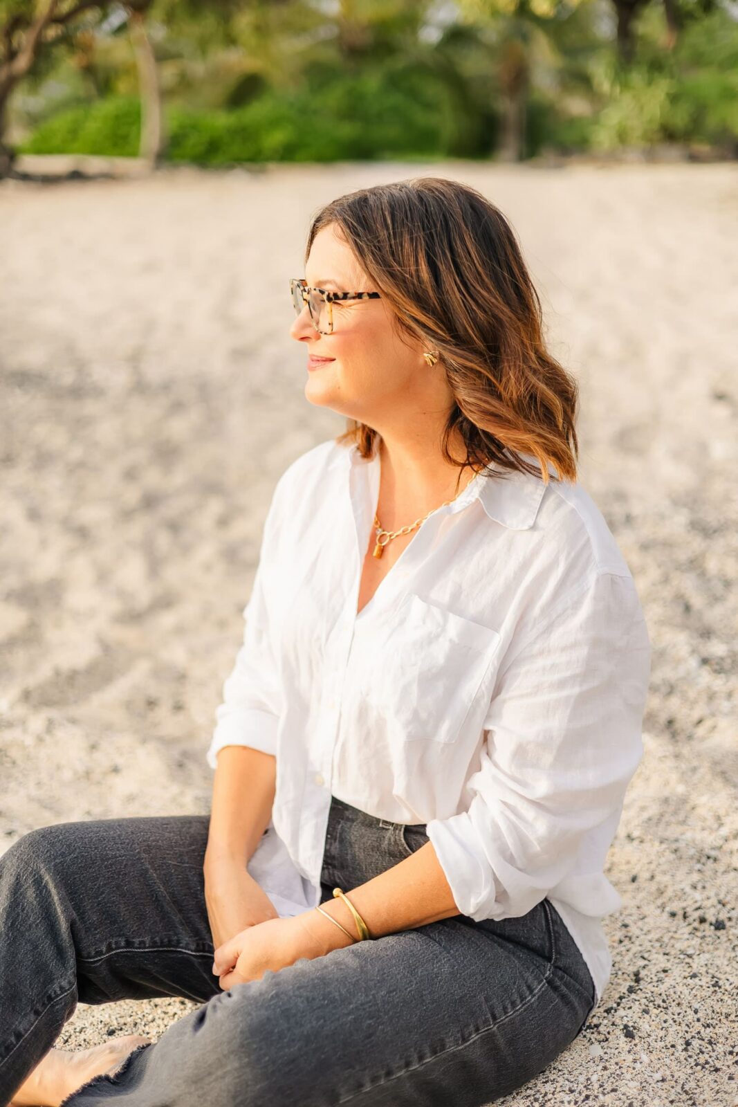 A woman with shoulder-length brown hair, wearing glasses, a white blouse, and dark jeans, sits on a sandy beach looking to the side. The background is blurred with greenery.