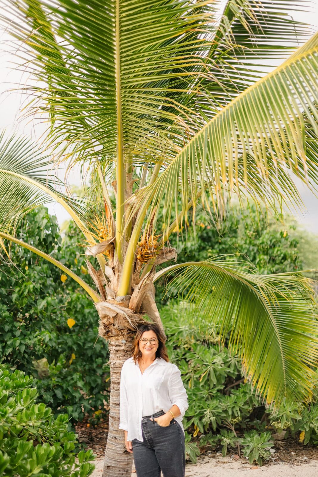 A woman wearing glasses, a white shirt, and dark pants stands smiling in front of a tall palm tree, surrounded by lush green foliage on a sunny day.