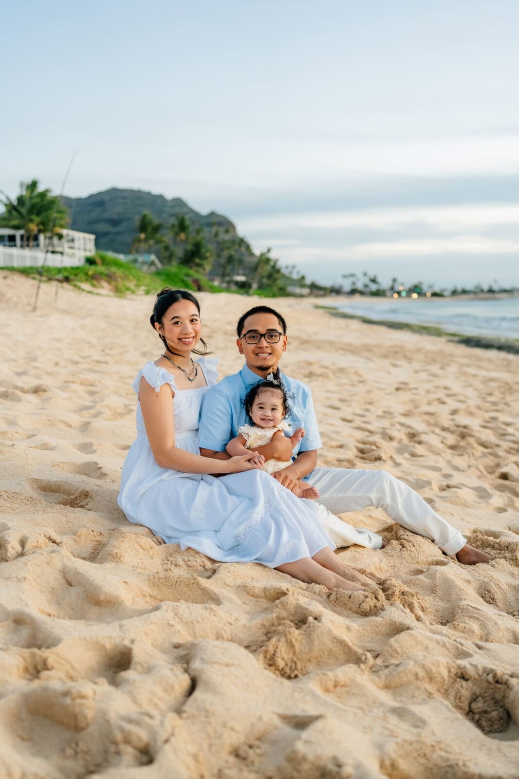 A family of three, dressed in light blue and white, sits on a sandy beach at sunset, smiling at the camera. Tropical greenery, houses, and mountains are visible in the background.