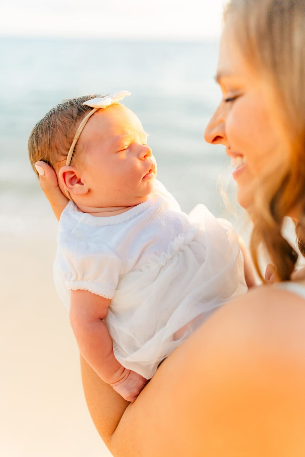 A woman smiling and holding a newborn baby dressed in white, with the sun shining on them and the ocean in the background.