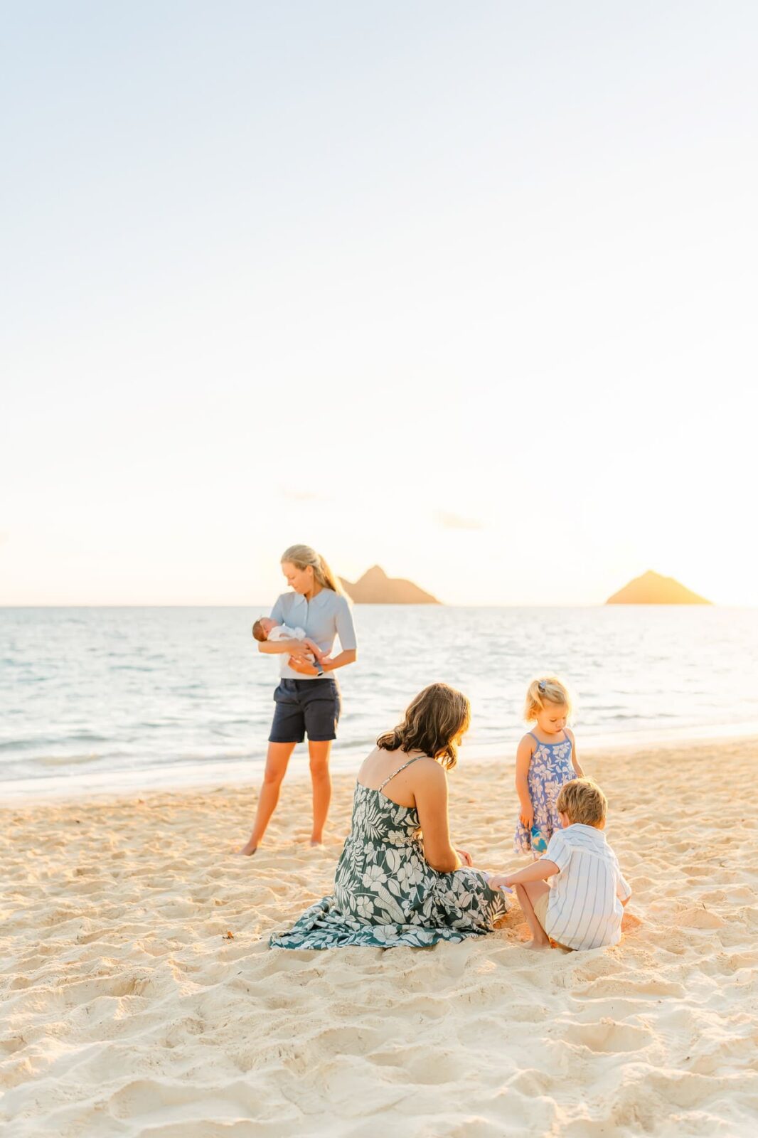 A woman in a floral dress sits on sandy beach with two young children, while another woman stands nearby holding a baby. The ocean and two small islands are visible in the background at sunset.