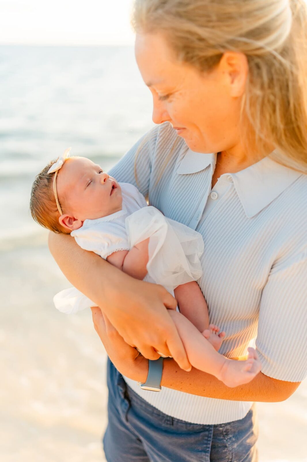 A woman smiles lovingly while holding a newborn baby dressed in white with a bow headband. They are outdoors near the water, and sunlight bathes the scene in a warm glow.