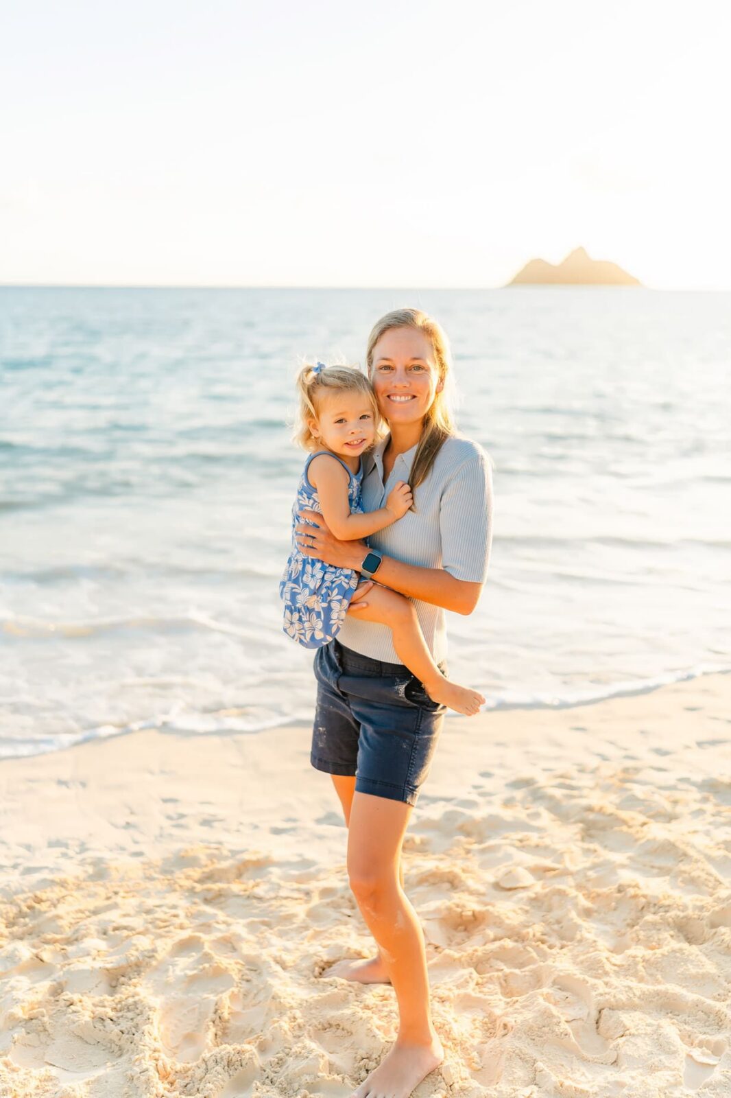 A woman stands barefoot on a sandy beach, smiling and holding a young girl. The ocean and distant islands are visible in the background under a bright, clear sky.