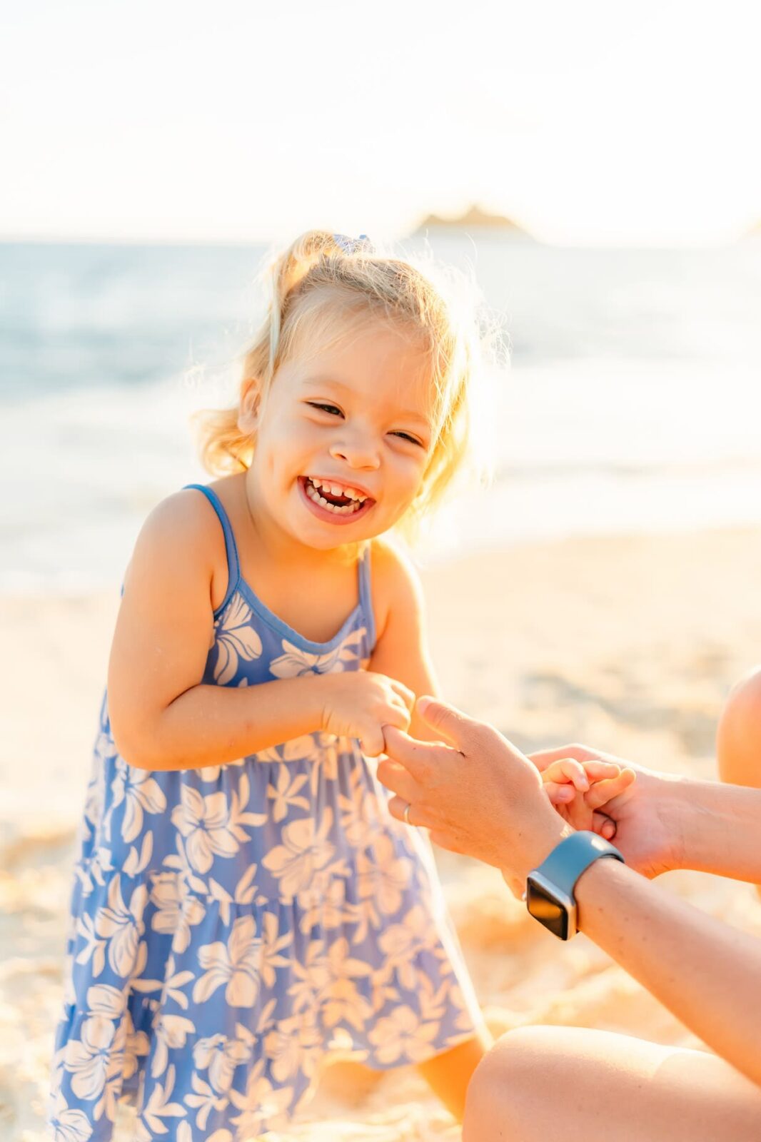 A smiling young girl in a blue floral dress stands on a sunny beach, laughing while holding hands with an adult whose arm wears a smartwatch. The ocean and a distant island are visible in the background.