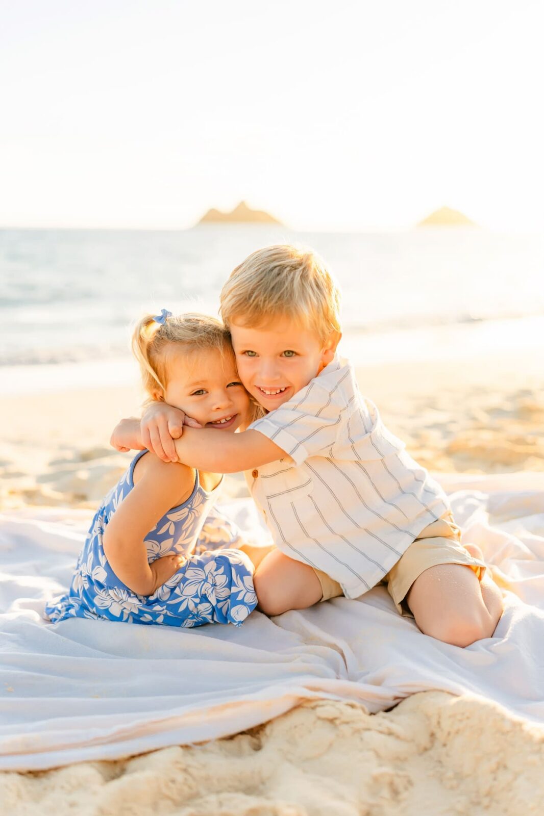 Two young children, a girl in a blue floral dress and a boy in a striped shirt, sit on a blanket at the beach, smiling and hugging each other with the ocean and islands in the background.