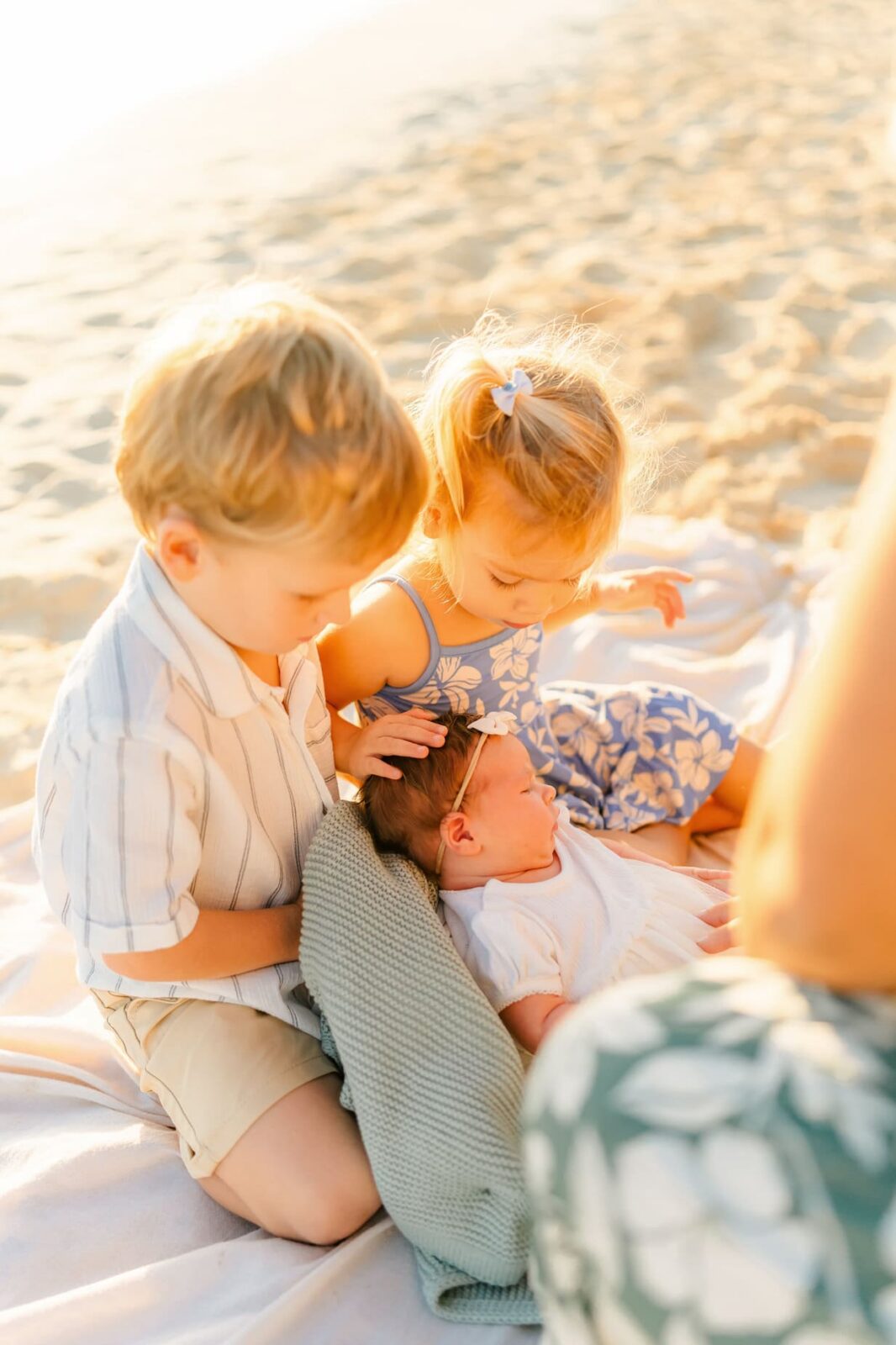 A young boy and girl gently touch a newborn baby lying on a blanket at the beach, bathed in warm sunlight. The boy wears a striped shirt and shorts; the girl wears a floral dress with her hair in a ponytail.