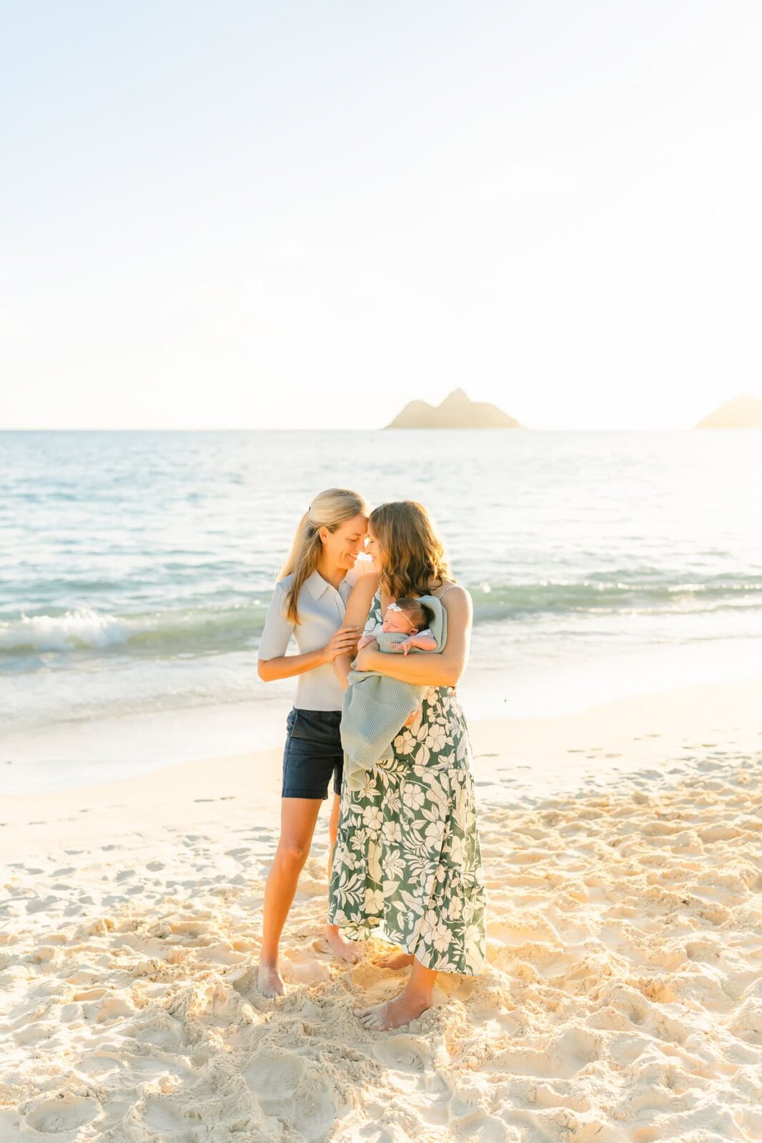 Two women stand on a sandy beach at sunset, one holding a baby wrapped in a blanket. The ocean and distant islands are in the background under a clear sky. All three are smiling and appear happy.