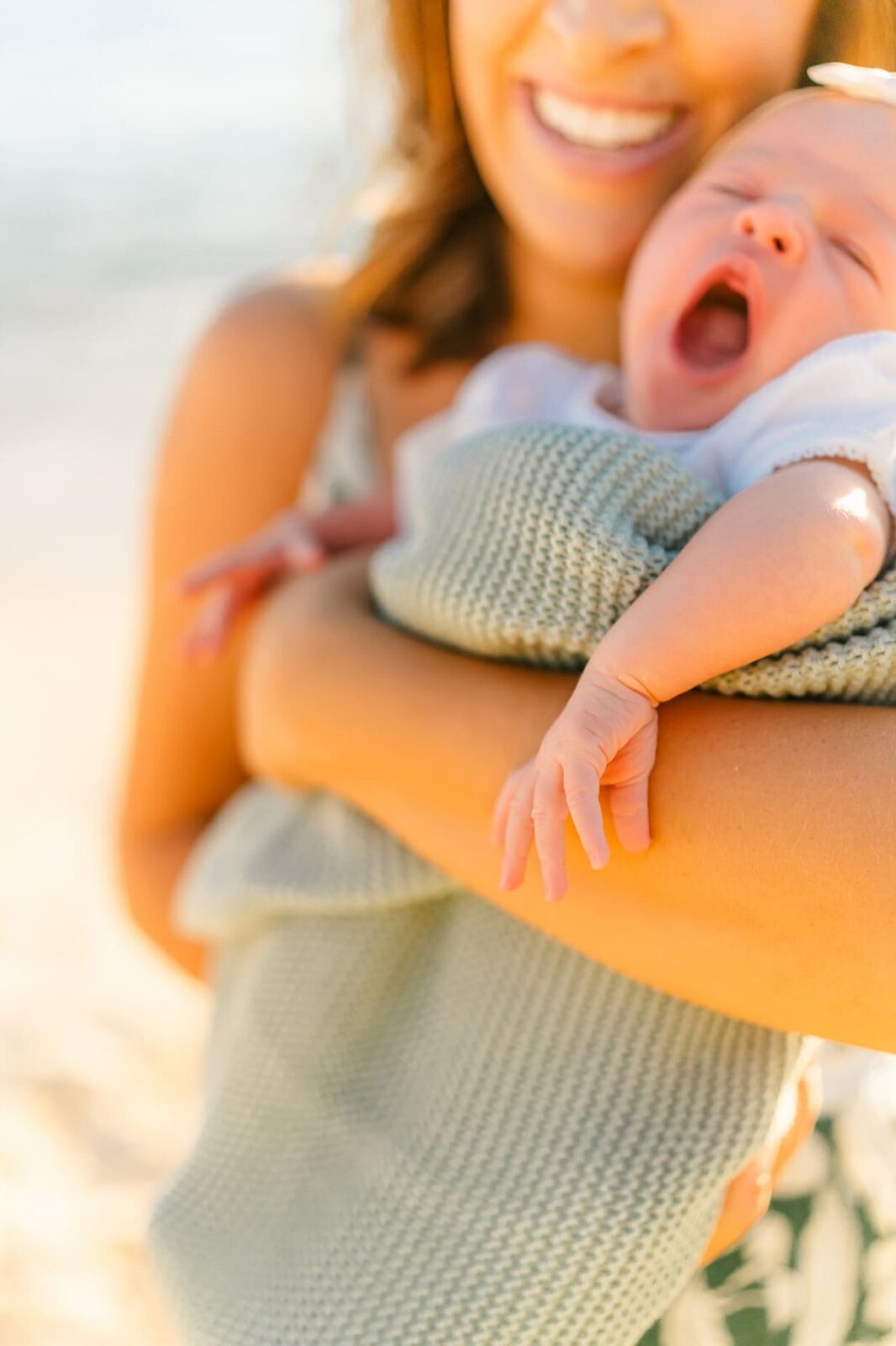 A woman smiles while holding a yawning baby wrapped in a green knitted blanket. The scene is bright and sunlit, suggesting a warm, outdoor setting.