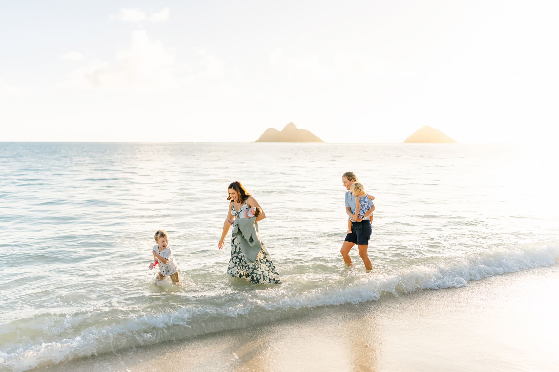Two adults and two young children walk and play in shallow ocean waves at a sunny beach, with two small islands visible in the background. The group appears joyful, enjoying a warm, bright day at the seaside.