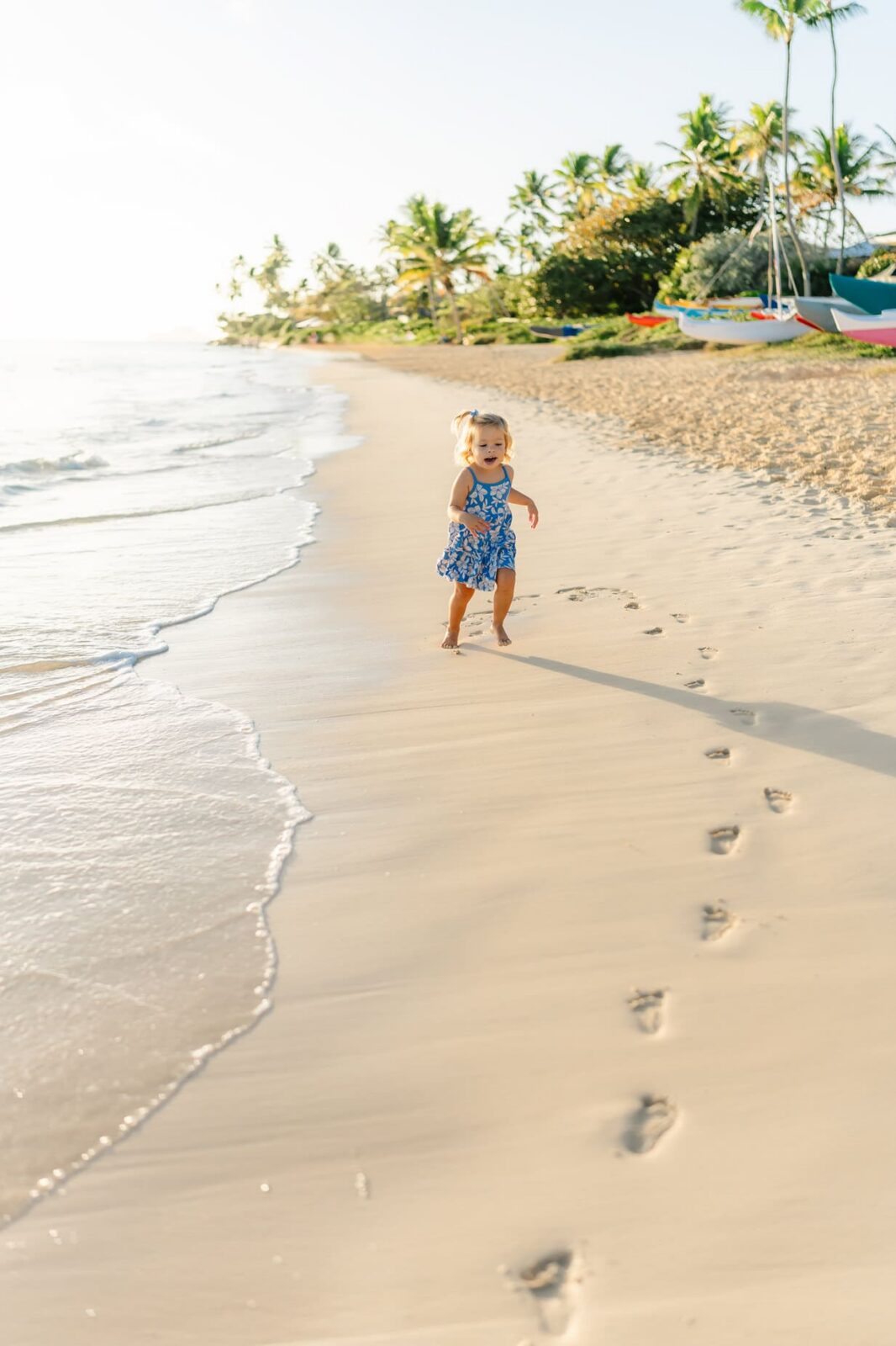 A young child in a blue dress walks along a sandy beach near the water, following a trail of footprints. Palm trees and boats are visible in the background under a clear sky.