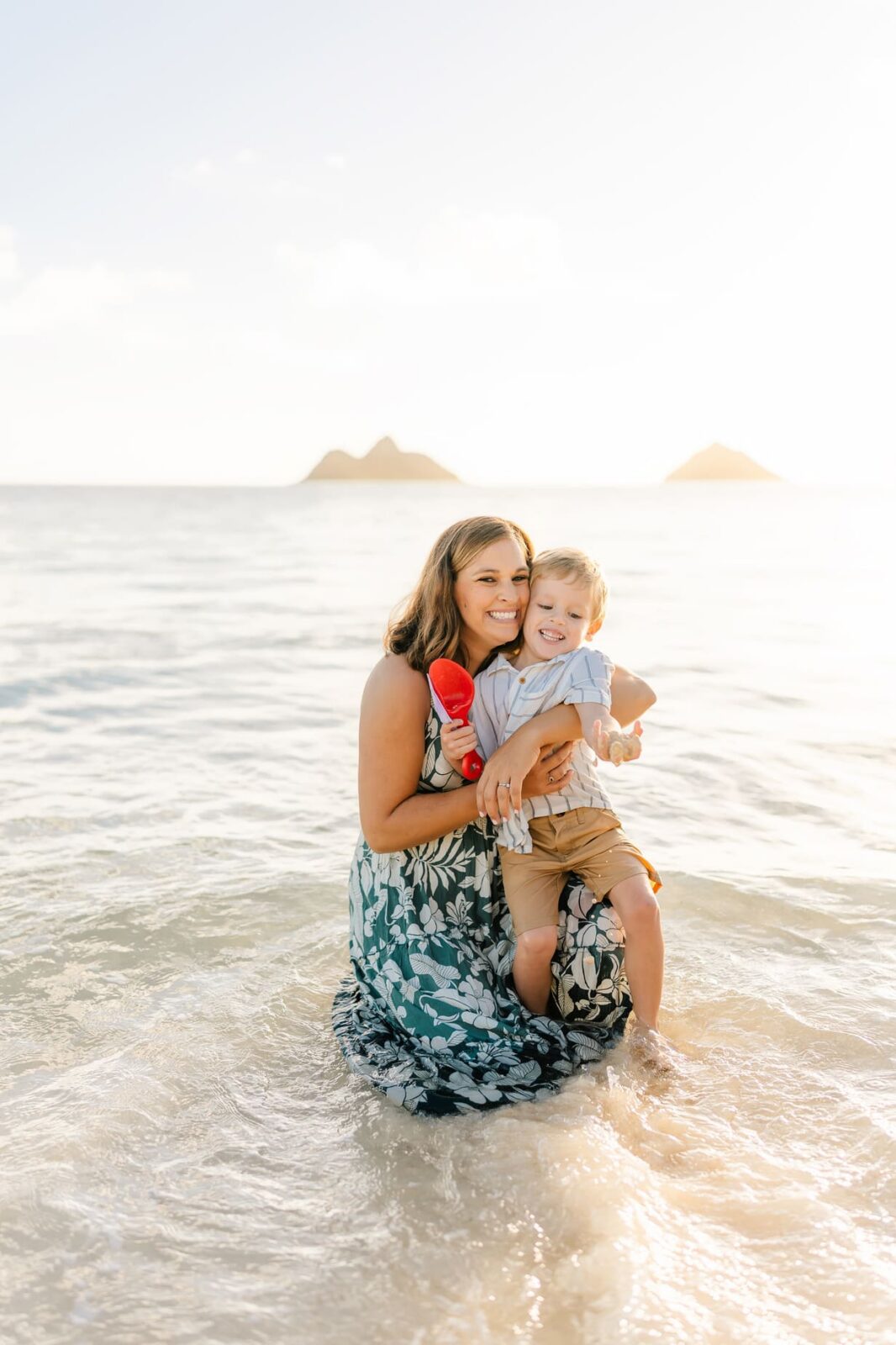 A woman in a green floral dress kneels in shallow ocean water, smiling and holding a young boy in her arms. The boy is laughing and holding a red toy, with two small islands in the background.